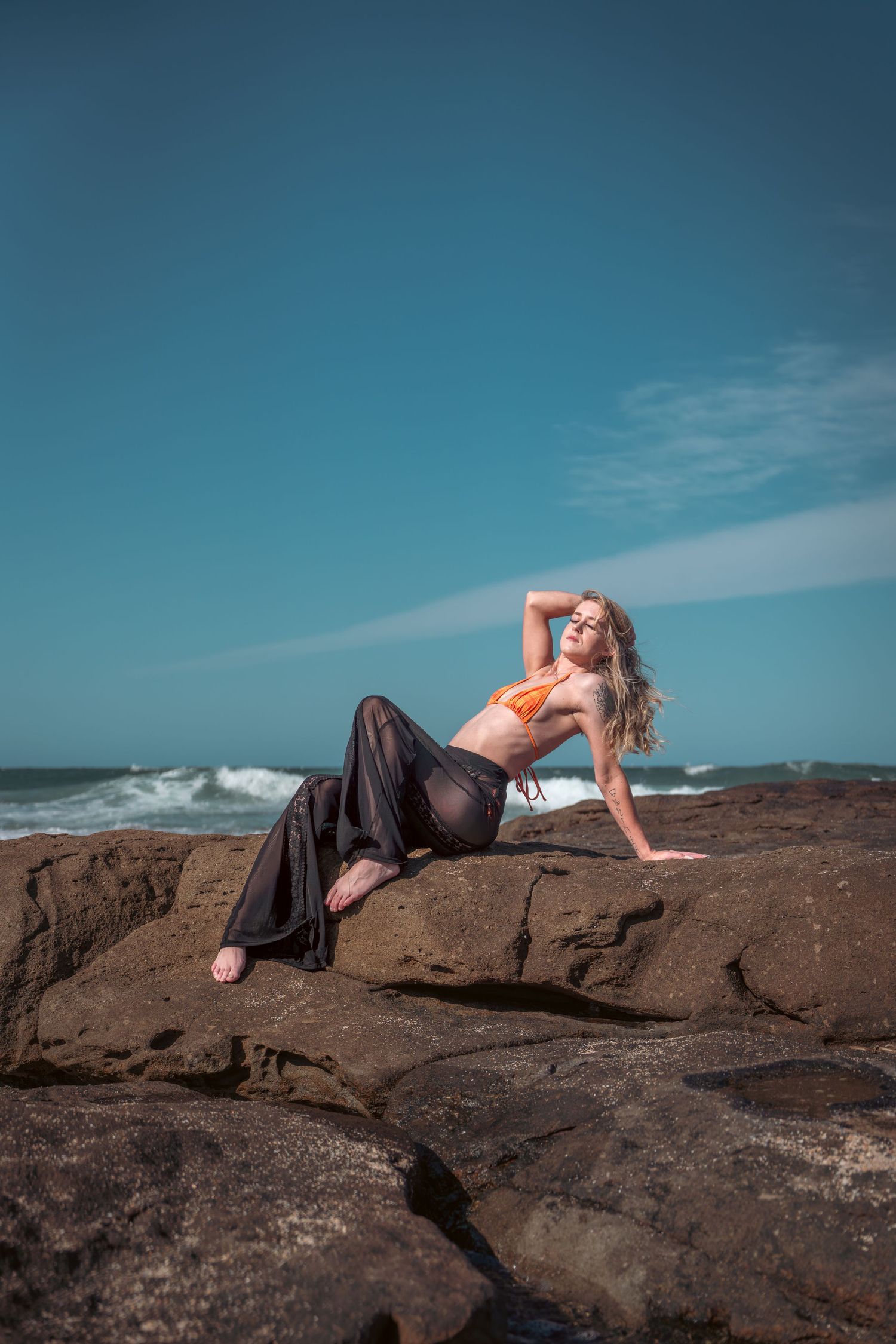 Model in flowing pants poses on coastal rocks against bright blue sky and ocean waves.