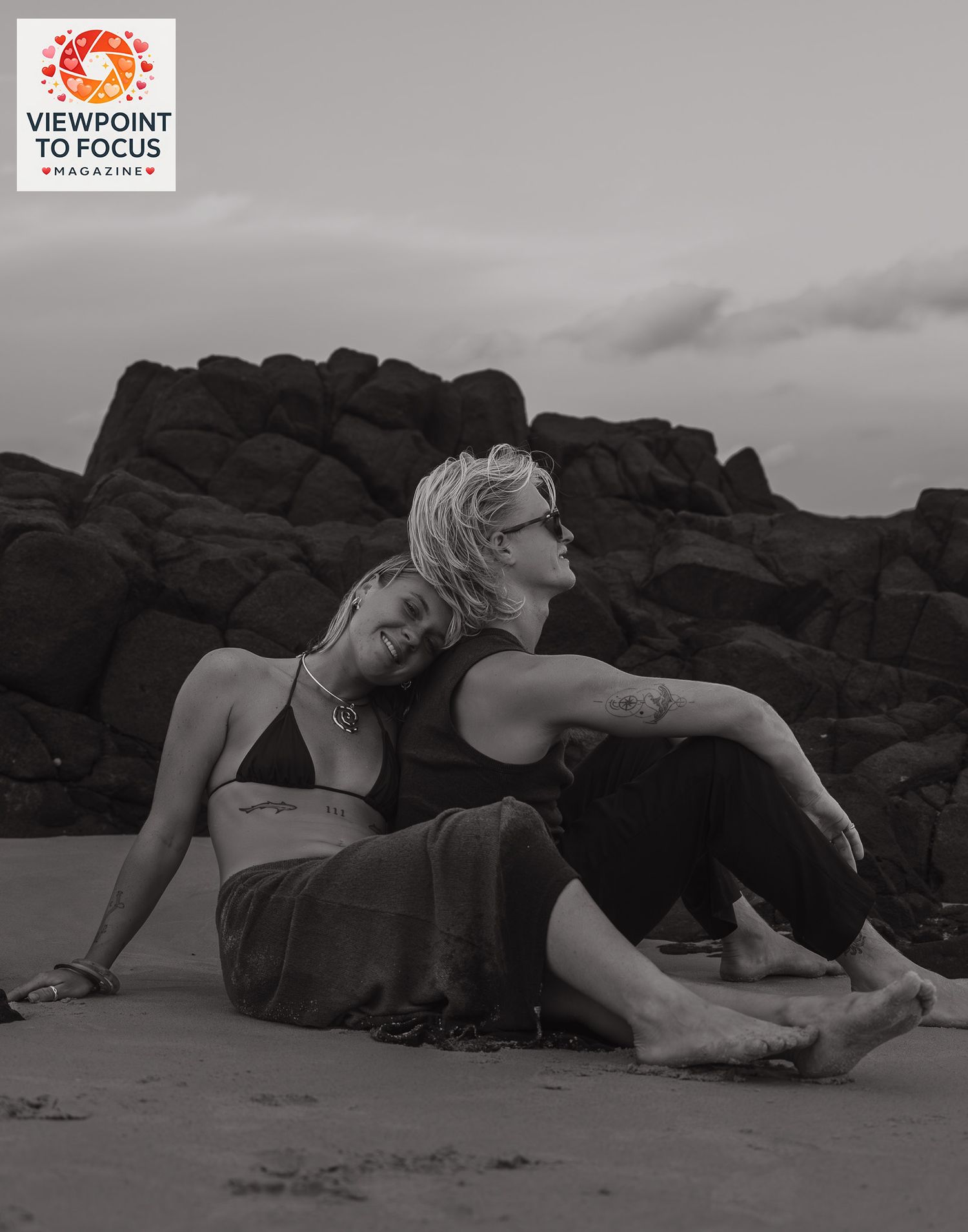 Two women sitting together on a sandy beach with rocky cliffs in black and white.