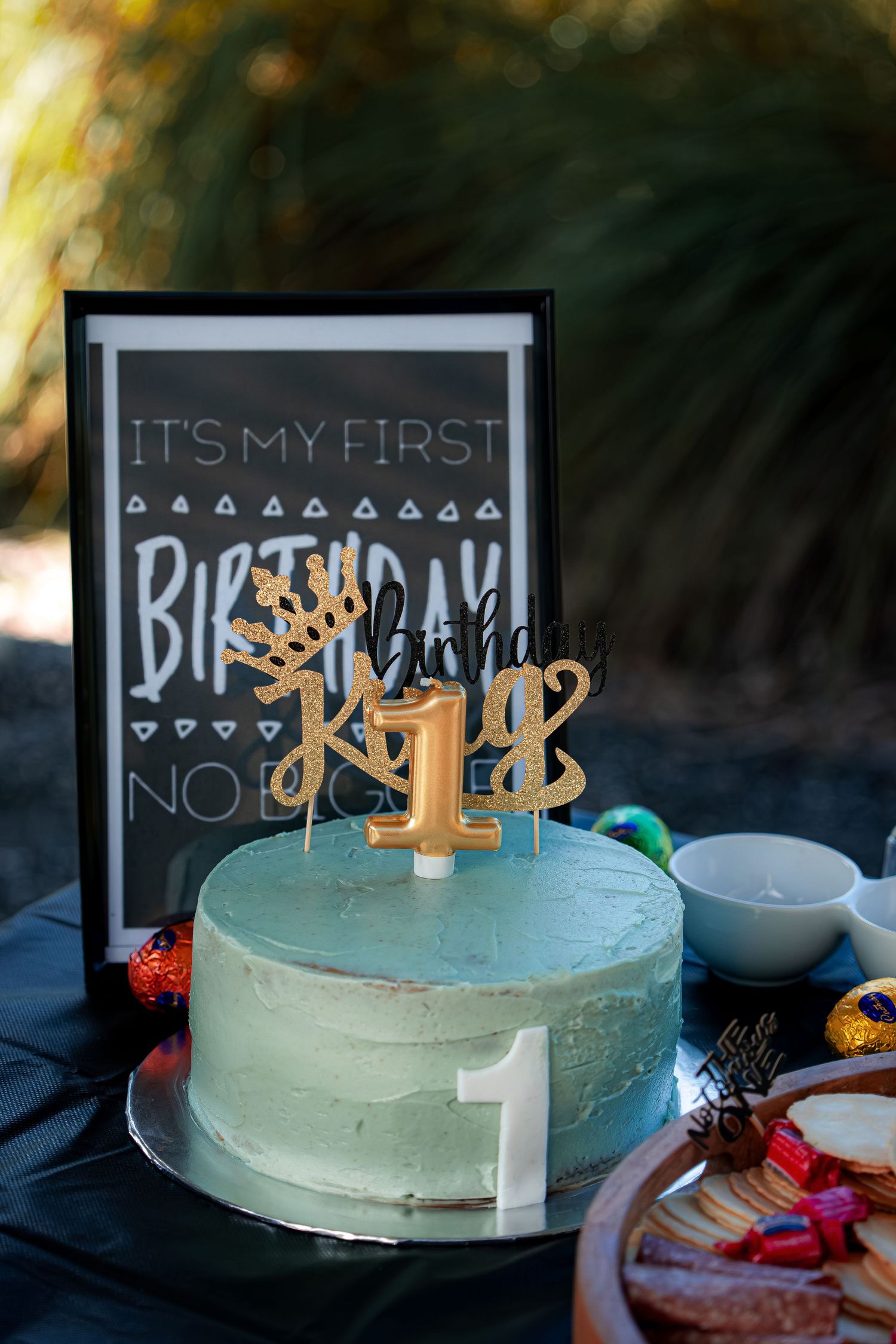 Light blue birthday cake with gold decorations and chalkboard signs displayed outdoors.