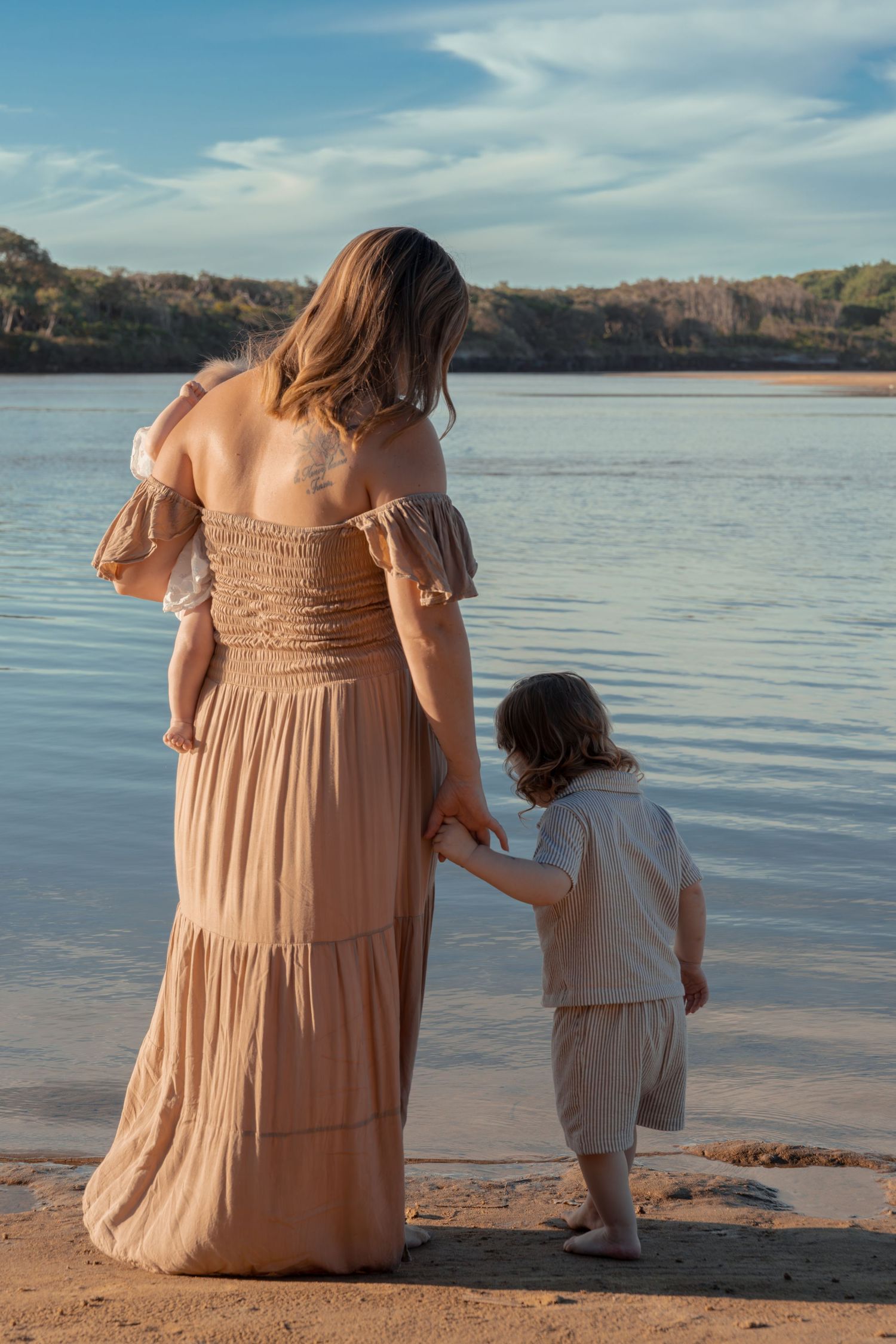 Woman in flowing dress stands with child at lakeside during sunset.