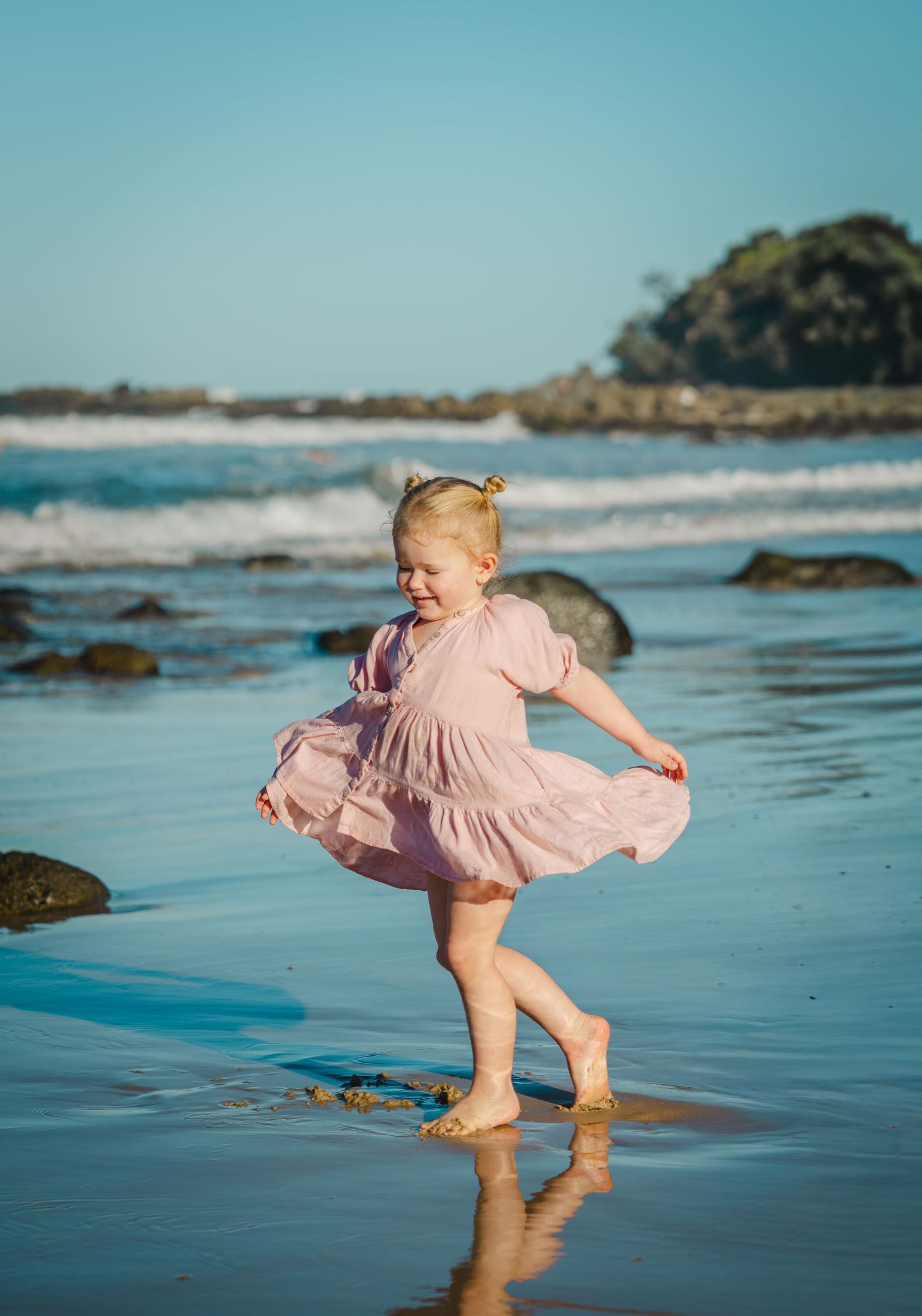 A joyful toddler girl in a pink dress twirls barefoot on a sunny beach with ocean waves in the background.