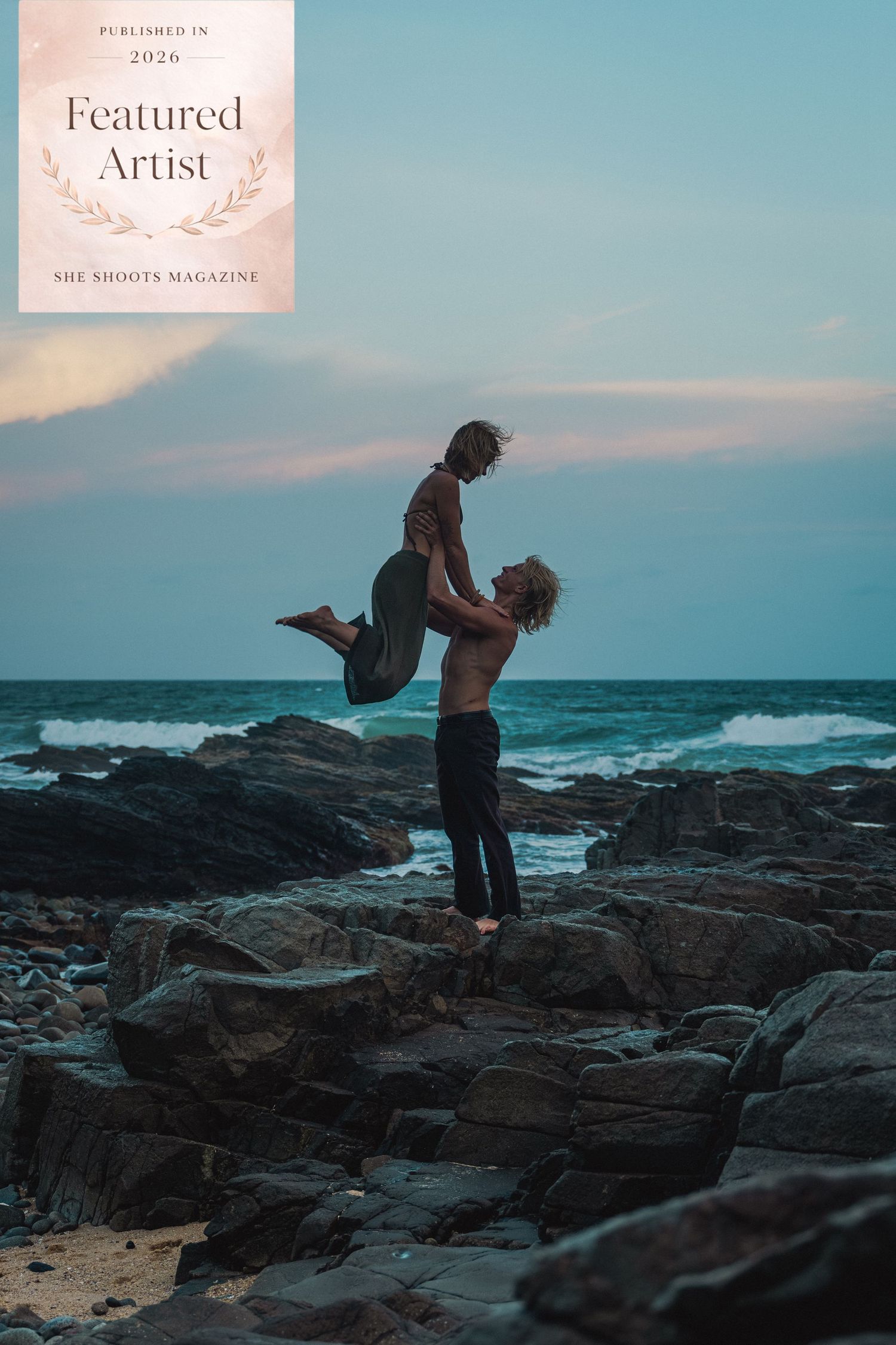 A man lifts a woman overhead on rocky seaside at dusk, featured in She Shoots Magazine 2026.