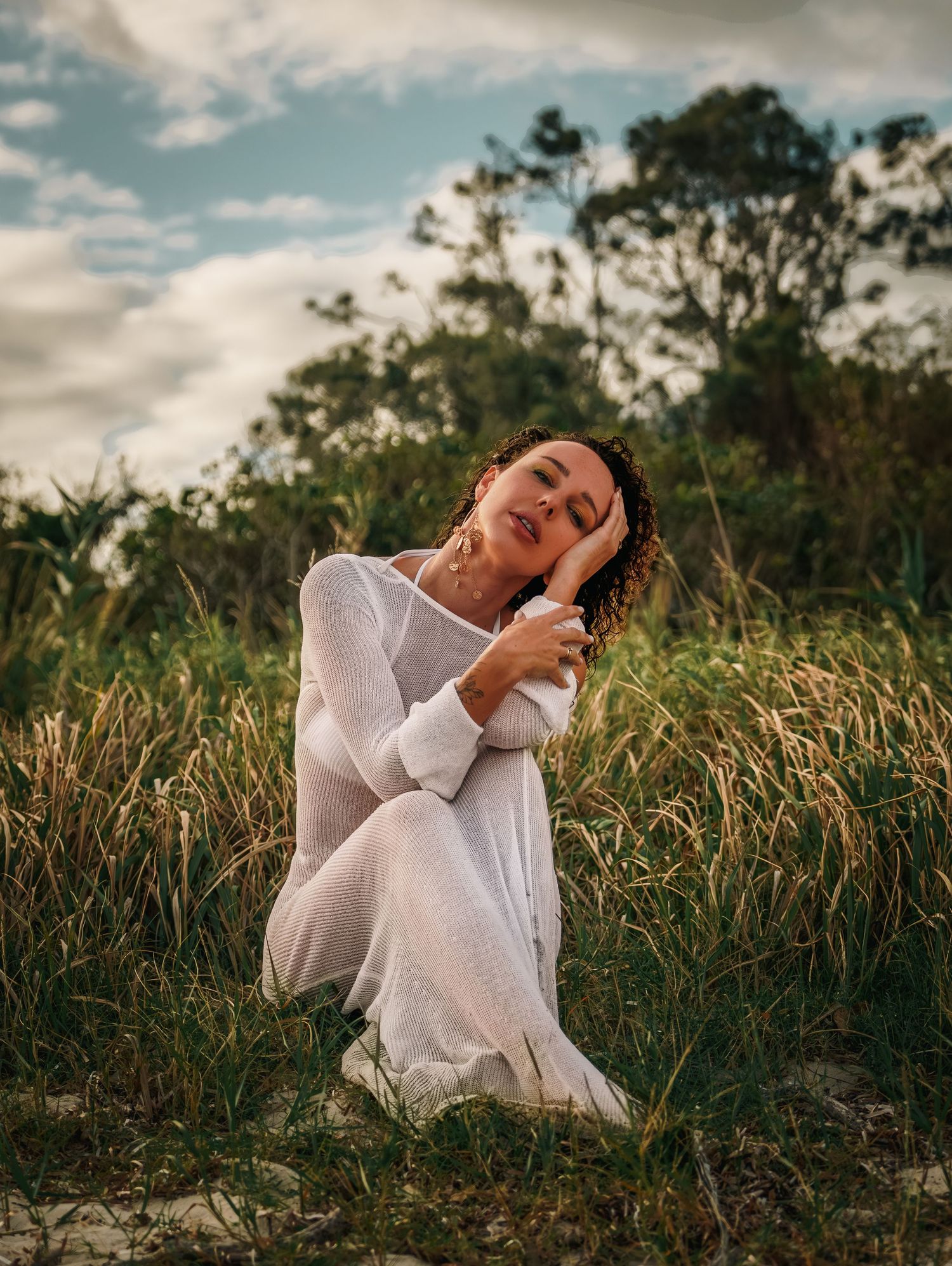 Woman in white dress kneeling in tall grass field, head tilted back, surrounded by lush green vegetation.