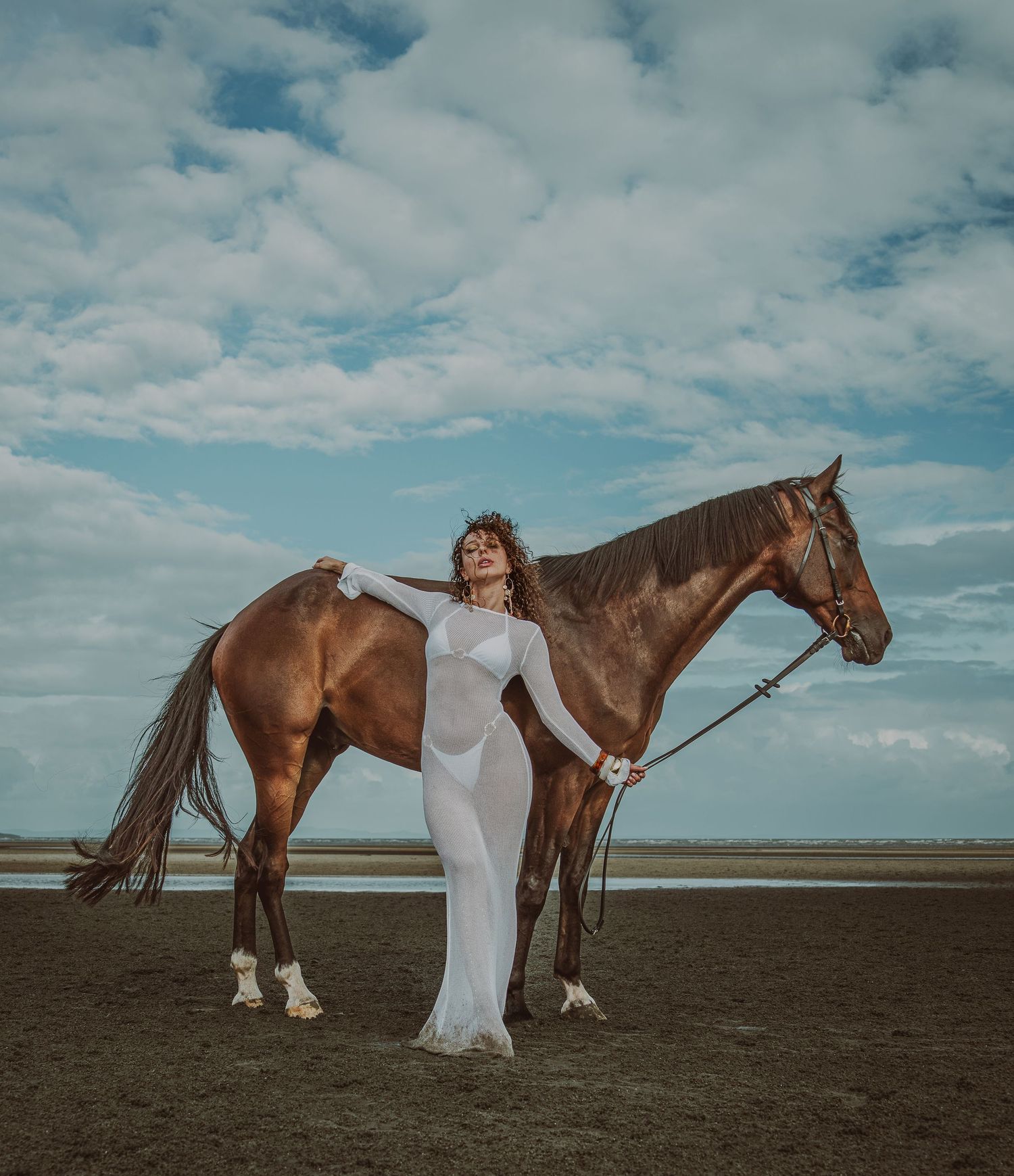 Woman in white gown poses with chestnut horse on dark sandy beach under dramatic cloudy sky.