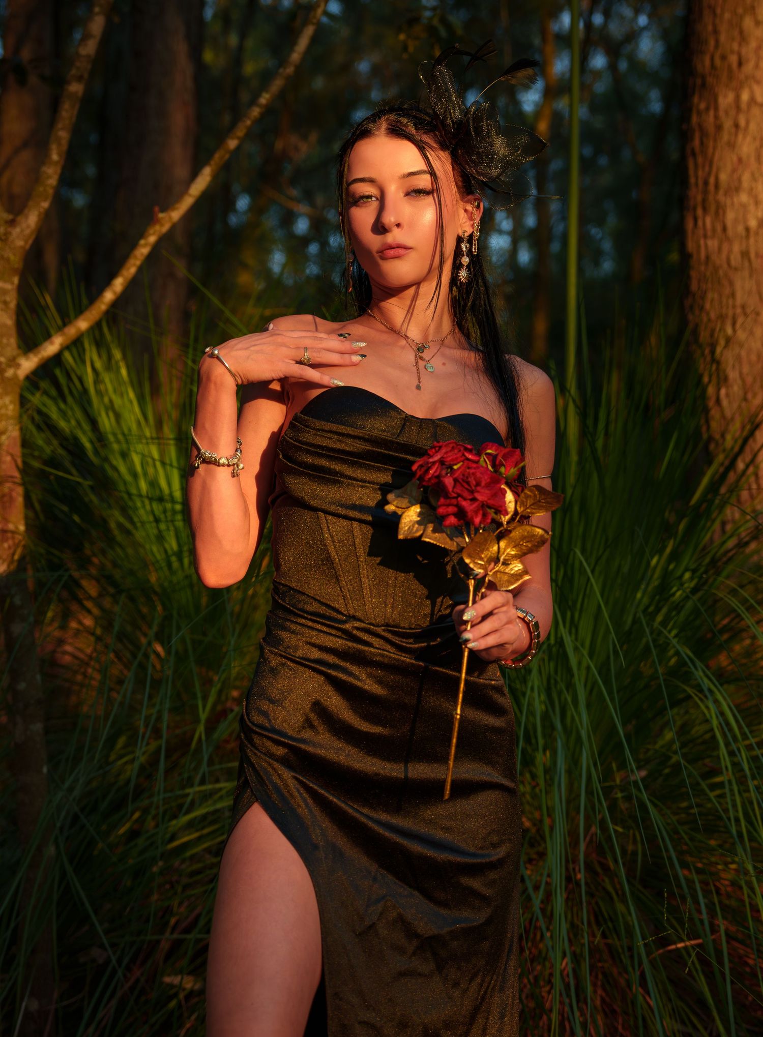Woman in black strapless dress holding red and gold roses poses in a sunlit forest setting.