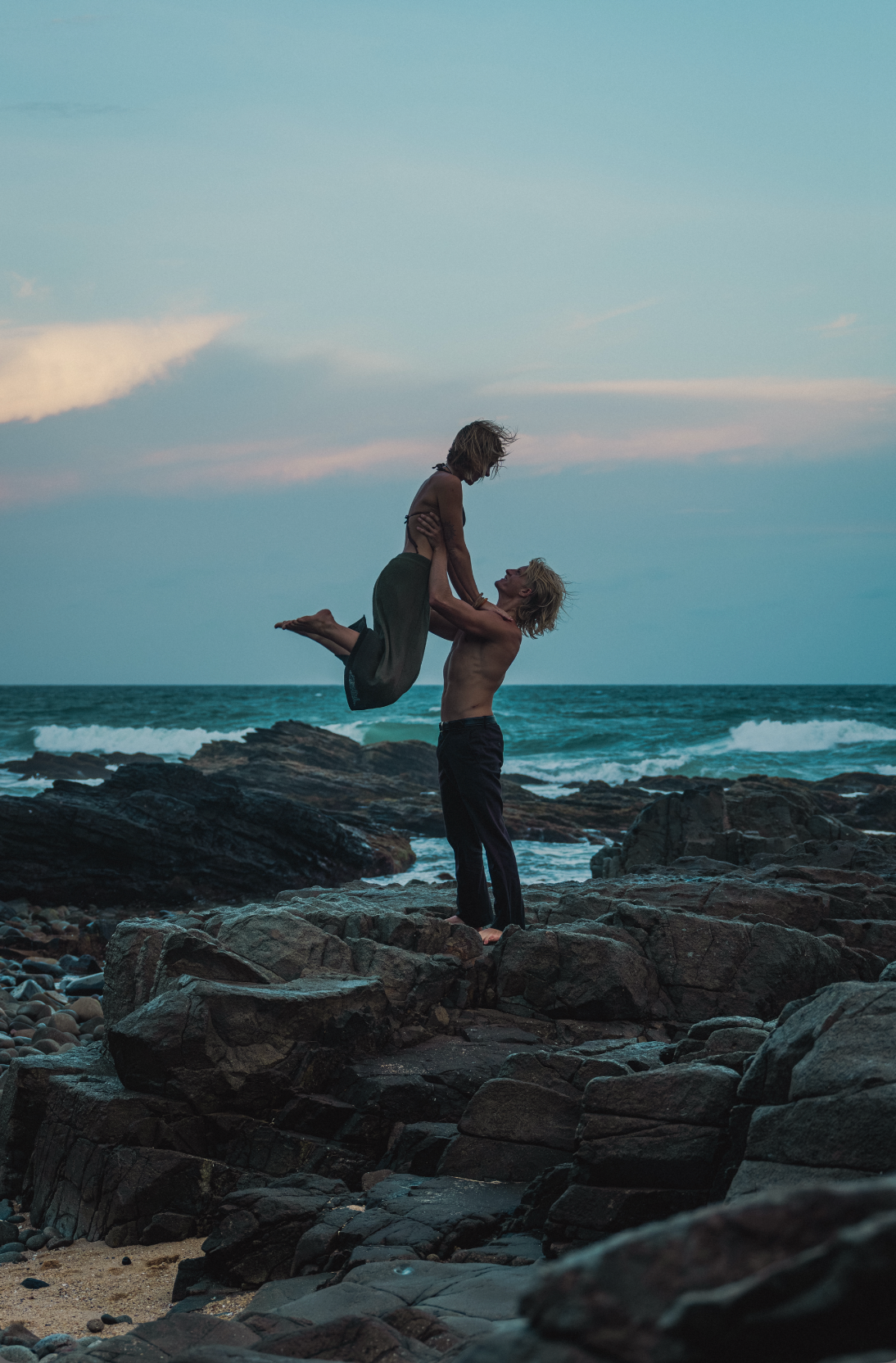 A shirtless man lifts a woman overhead on rocky coastal shore at dusk with turquoise ocean waves behind them.
