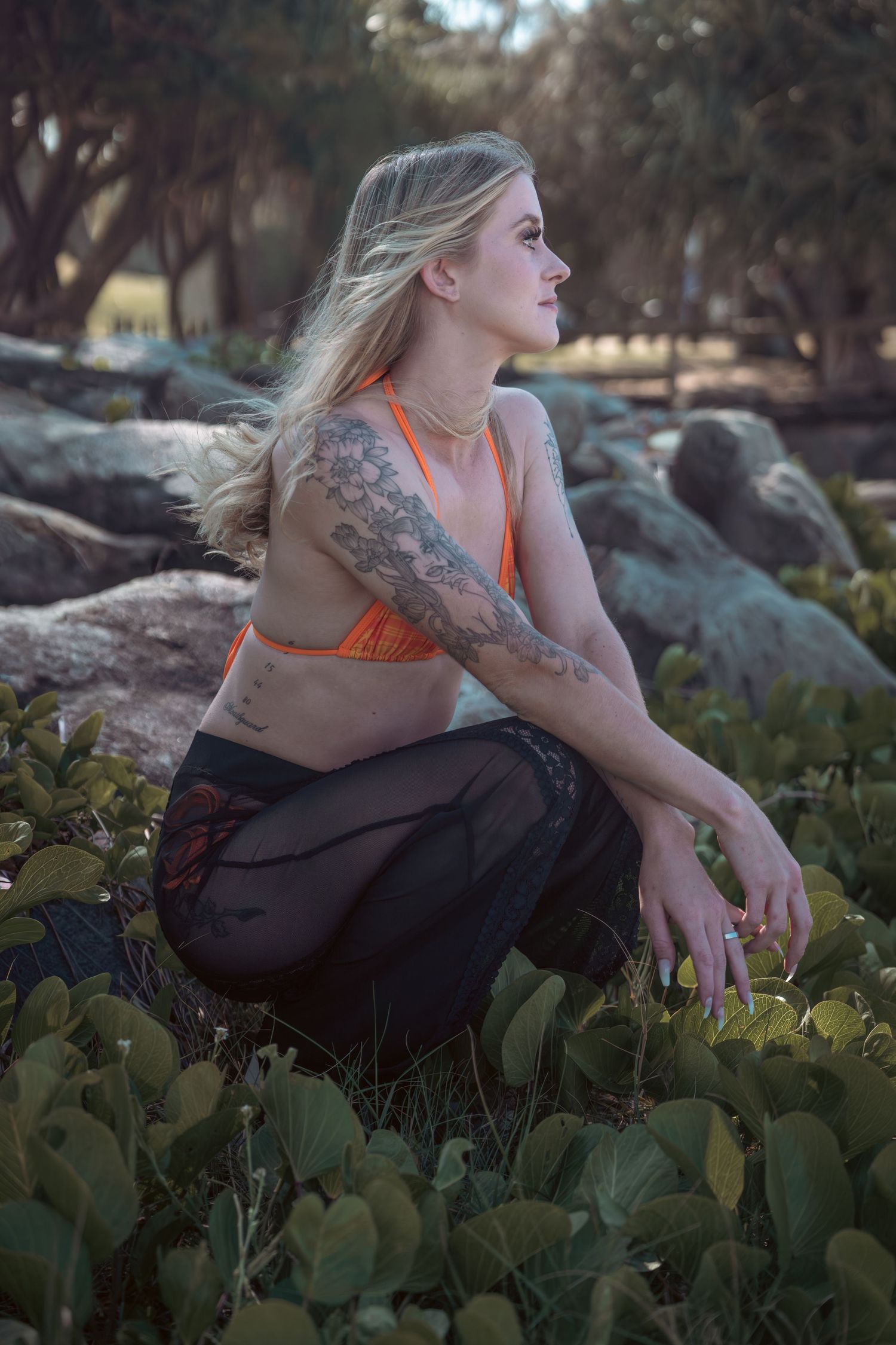 Someone in orange top and dark pants poses thoughtfully among rocks and foliage.