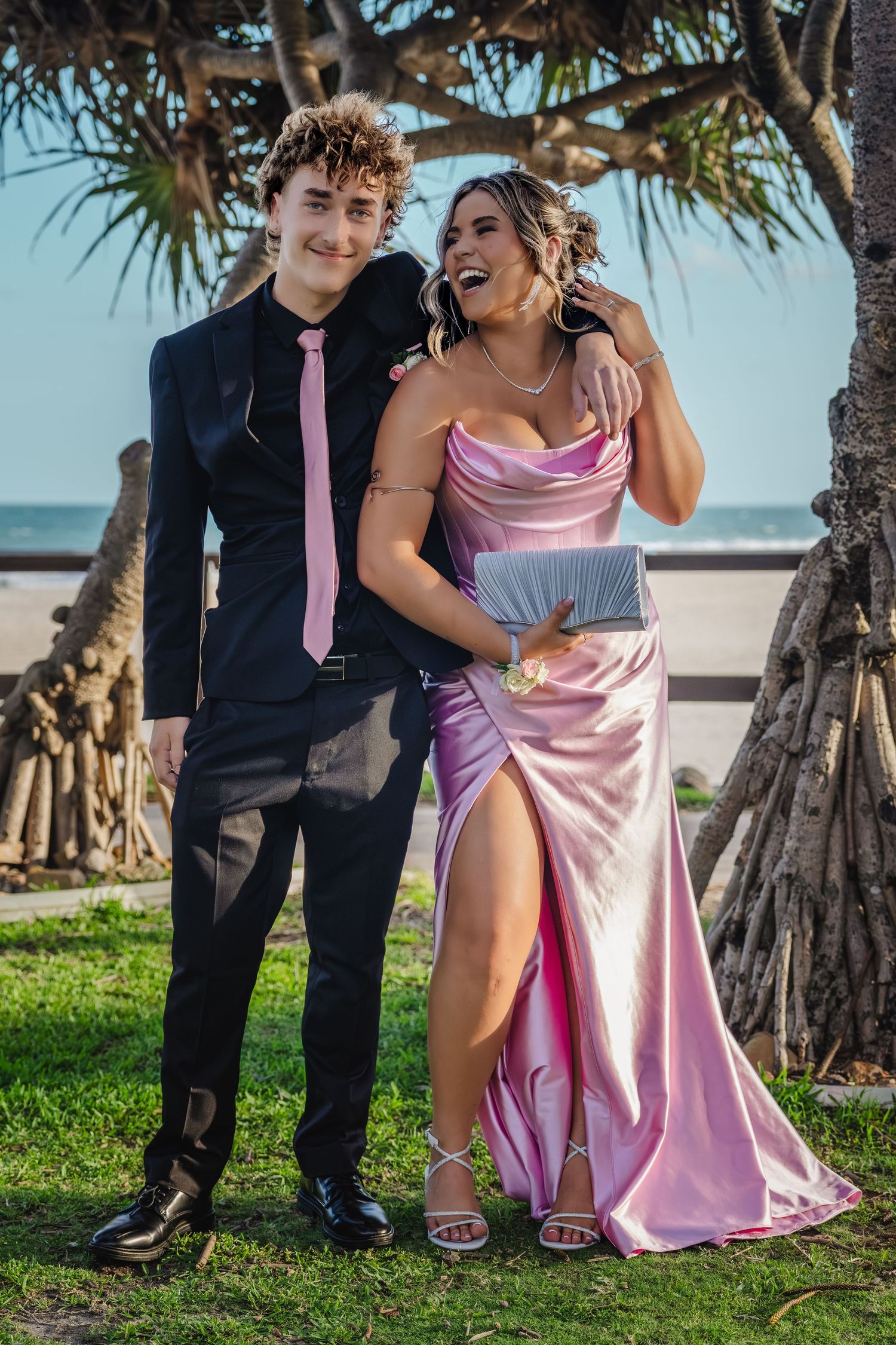Two people in contrasting formal wear share laughs while posing on grass near a beach.