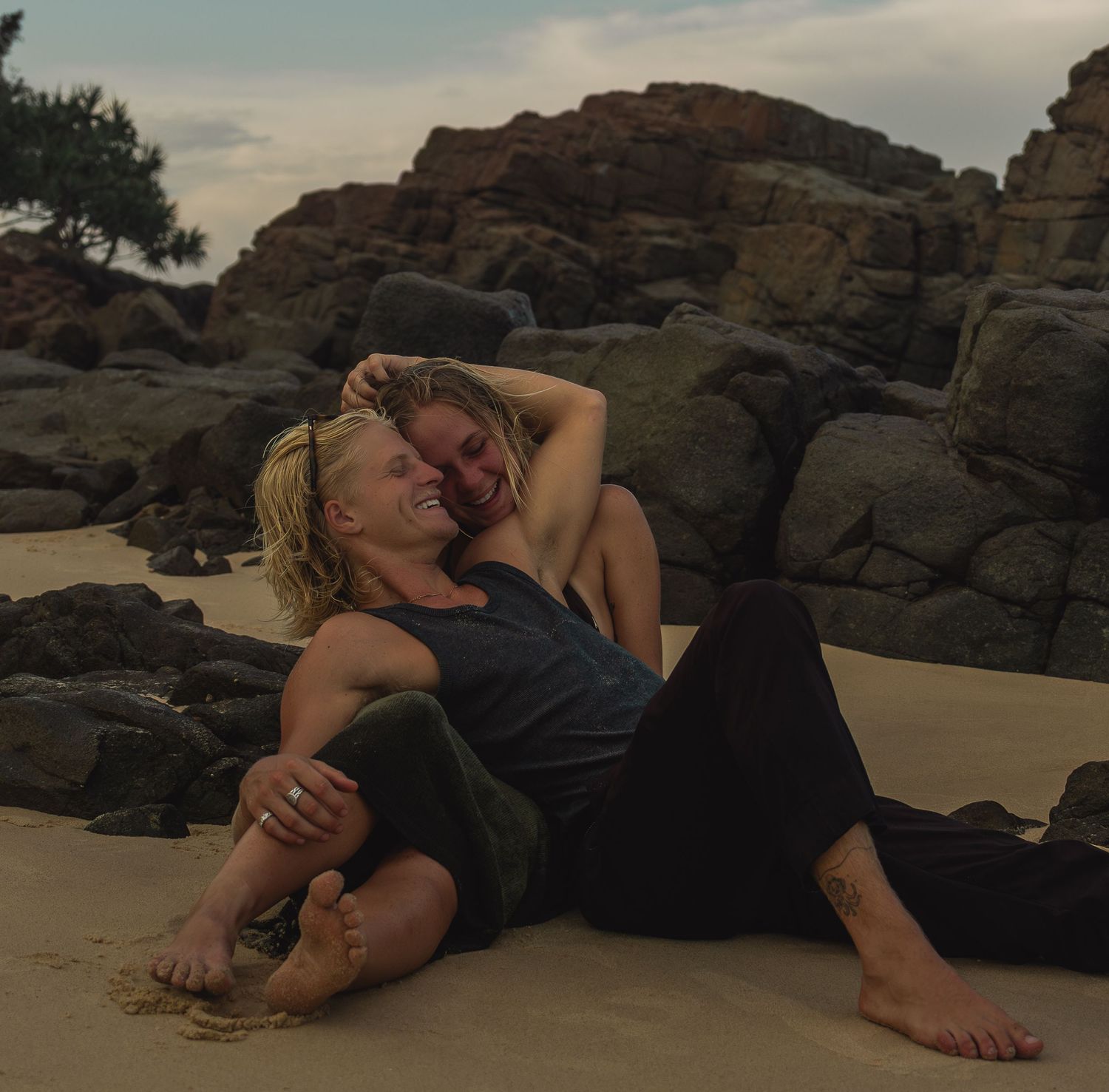 Two people relaxing on a sandy beach with dramatic rock formations in the background at sunset.