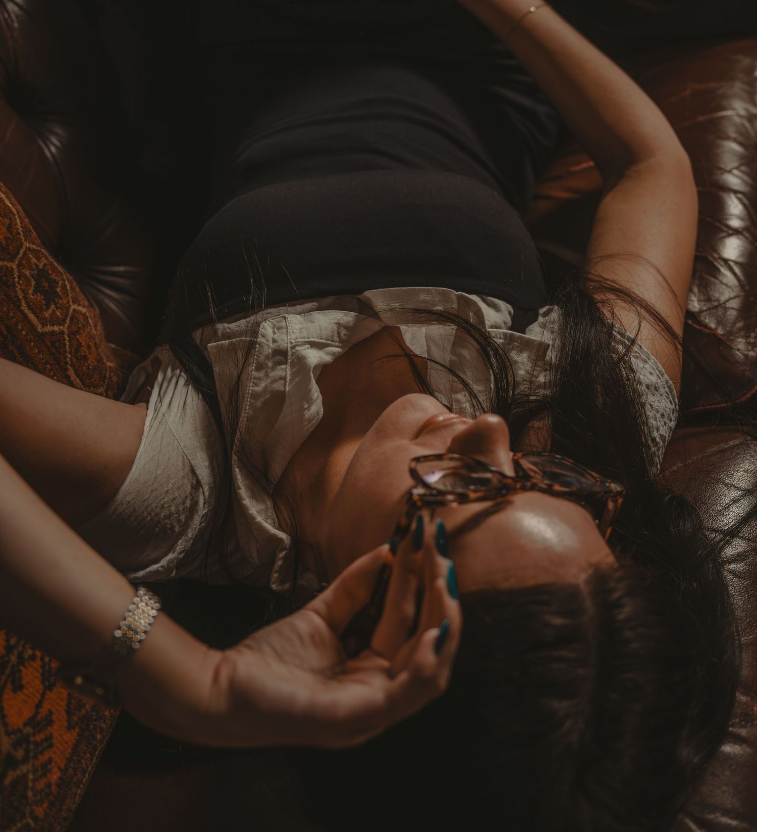 Woman reclining on leather sofa in black top and shorts, holding sunglasses with long dark hair.