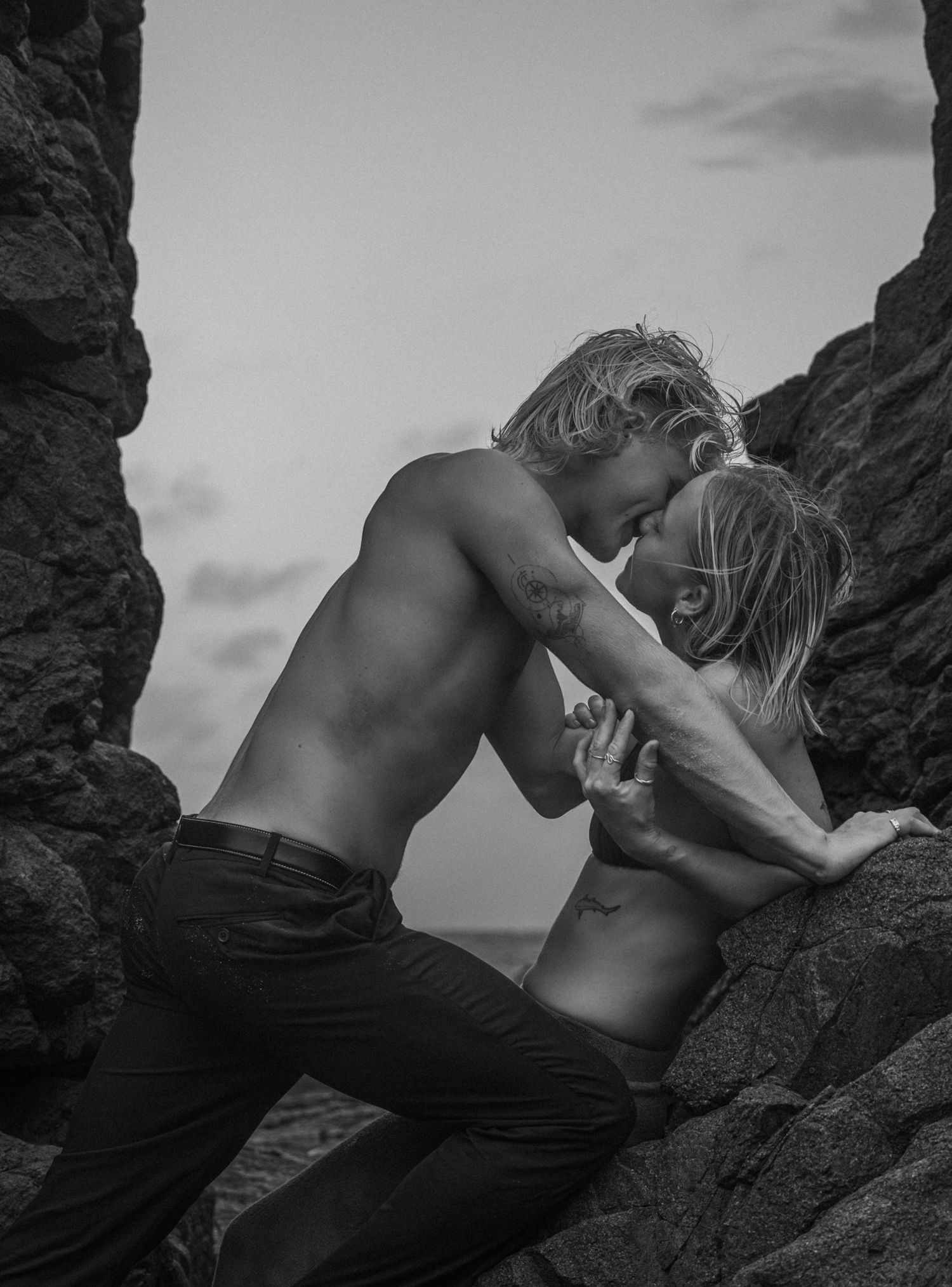 Black and white photo of a shirtless couple sharing an intimate moment among rocky coastal formations.