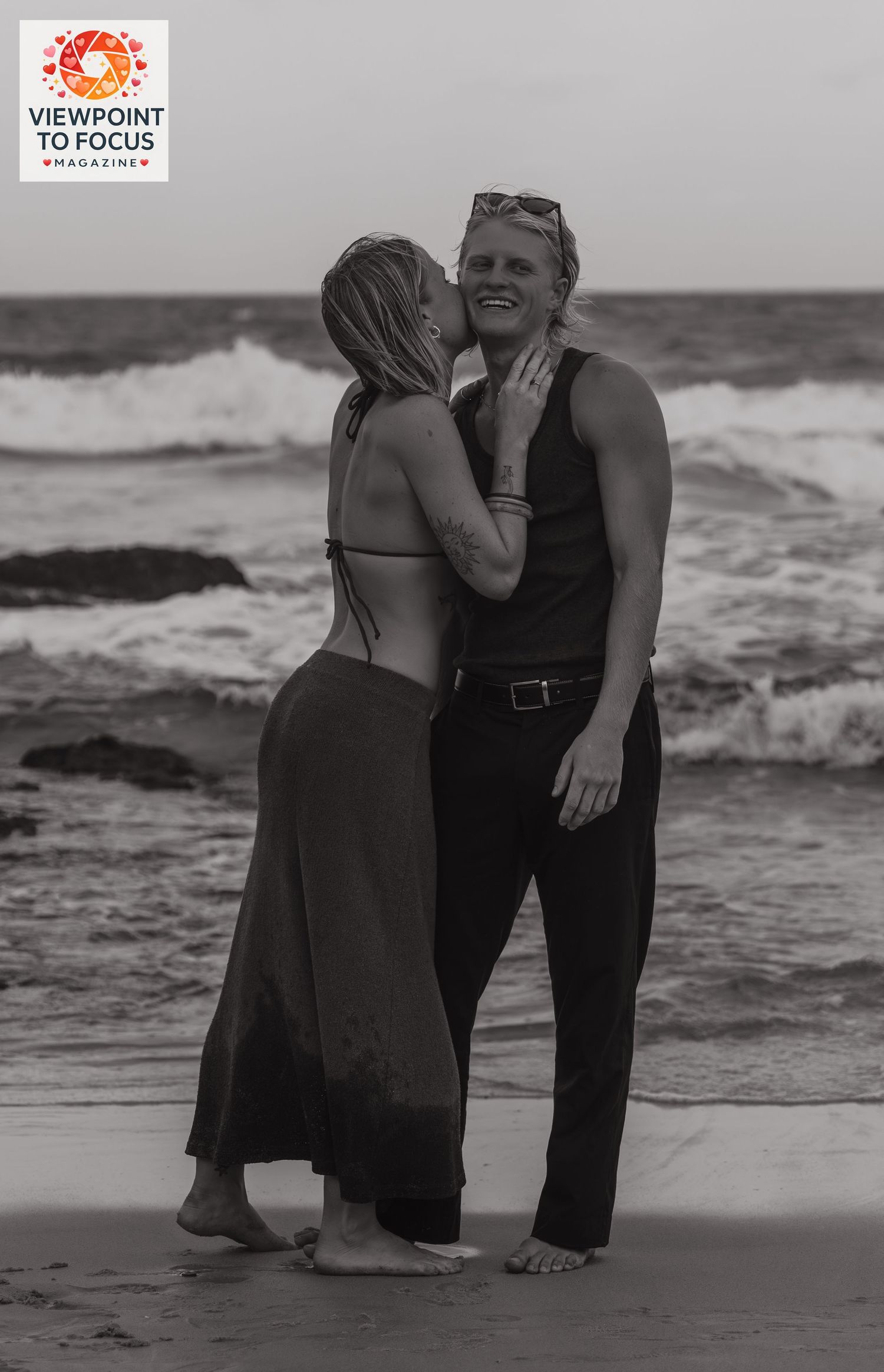 Couple sharing a romantic moment on a beach, woman kissing mans cheek while waves crash behind them.