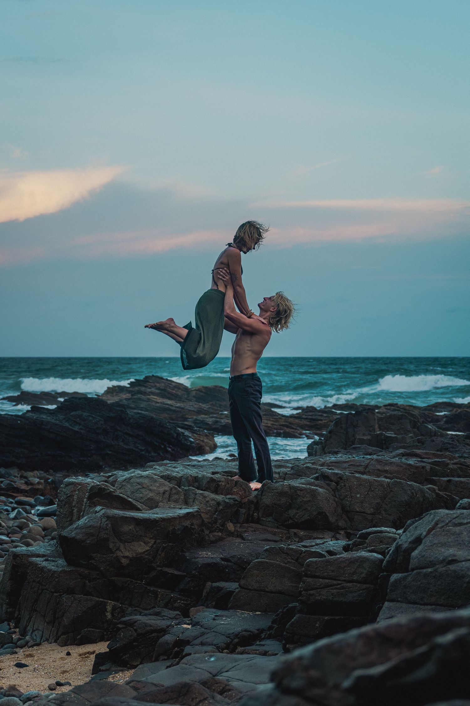 A silhouetted couple performs an acrobatic lift pose on rocky coastal shores at sunset against a turquoise sky.