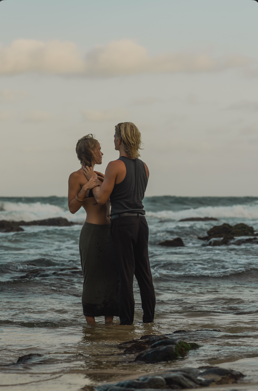 Couple standing together in ocean waves at rocky beach during golden hour, woman in black skirt embracing man in dark tank top.
