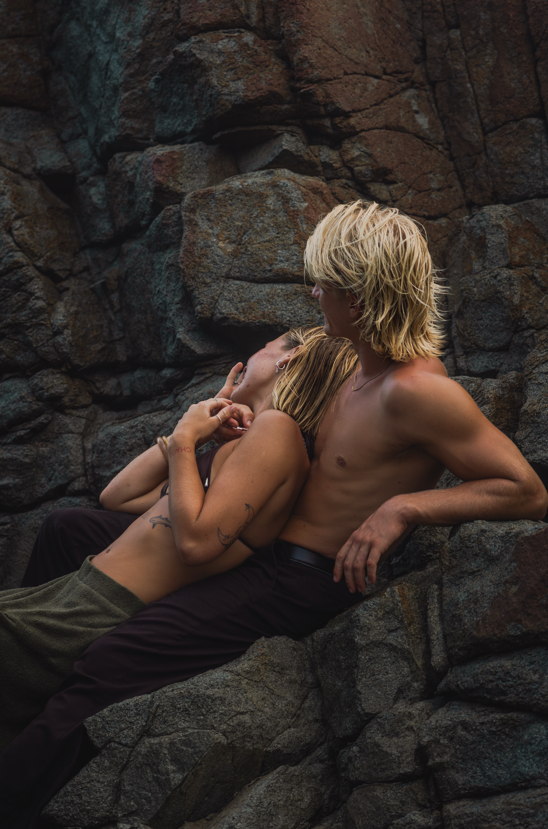 Sunshine Coast Photographer A shirtless blond man and woman embrace romantically against rugged rocky cliffs in an outdoor setting.