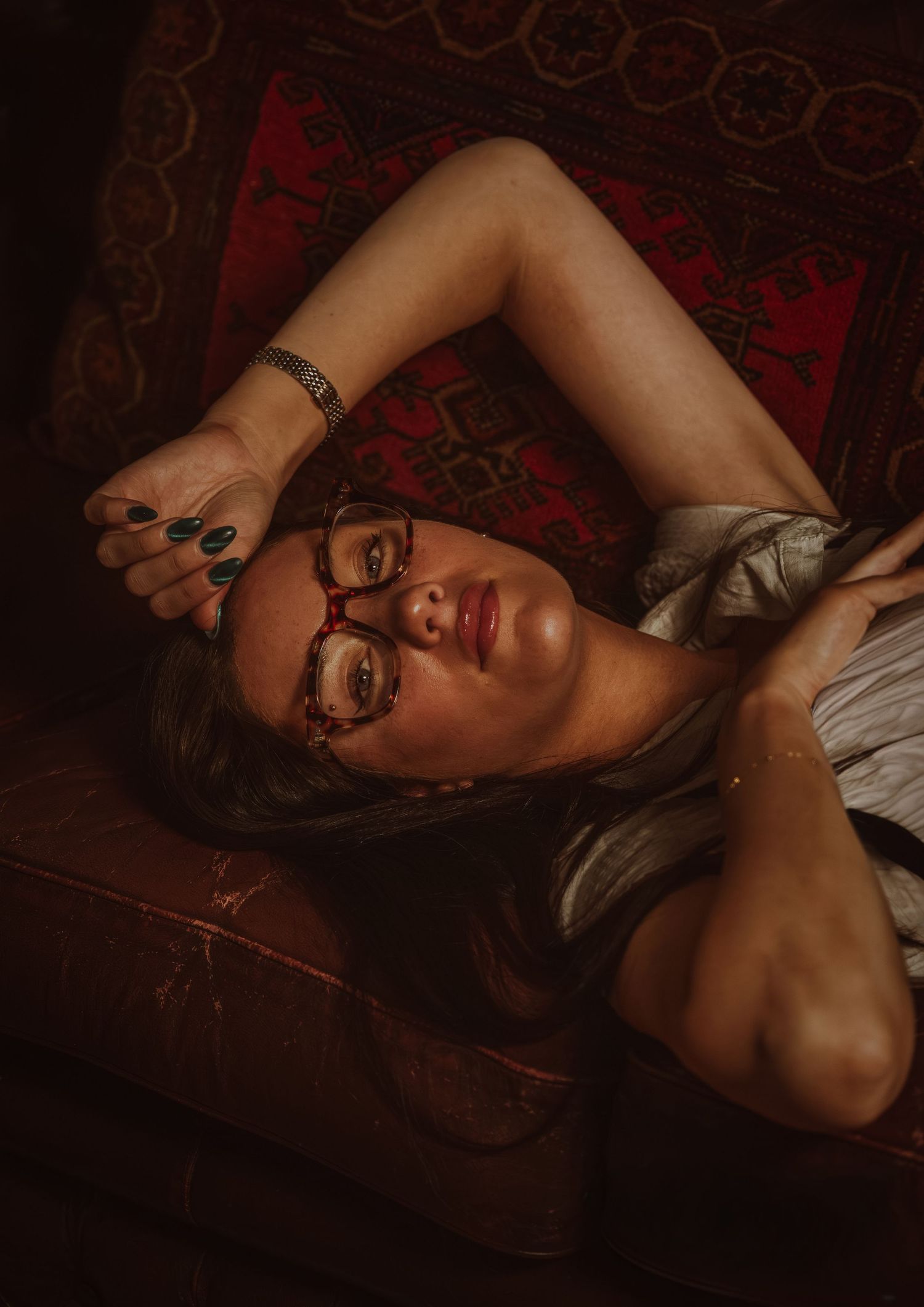 Woman with glasses and dark nails reclines on vintage furniture with red patterned upholstery in moody lighting.