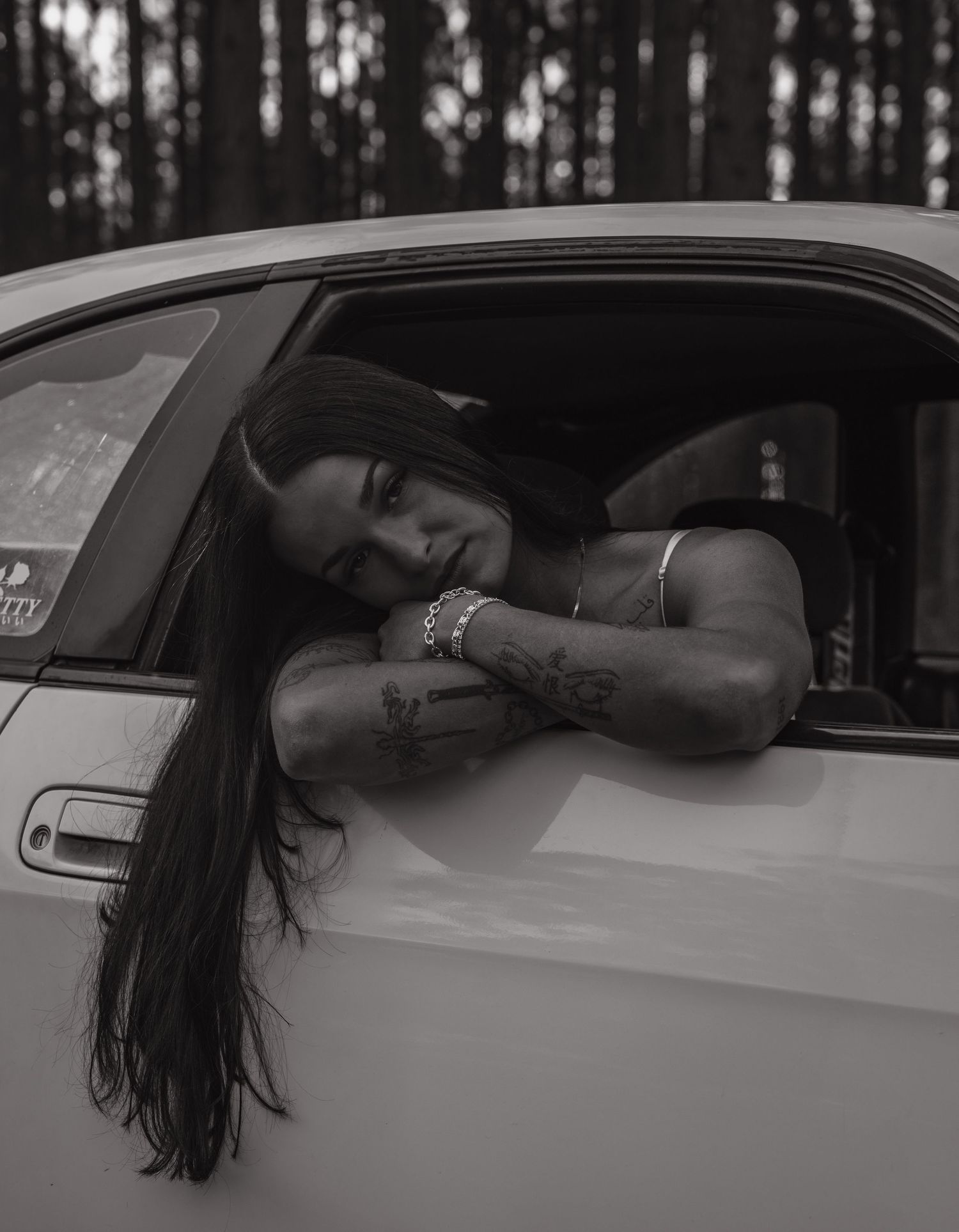 Black and white photo of a tattooed woman leaning out of a white car window, long dark hair, bracelets.