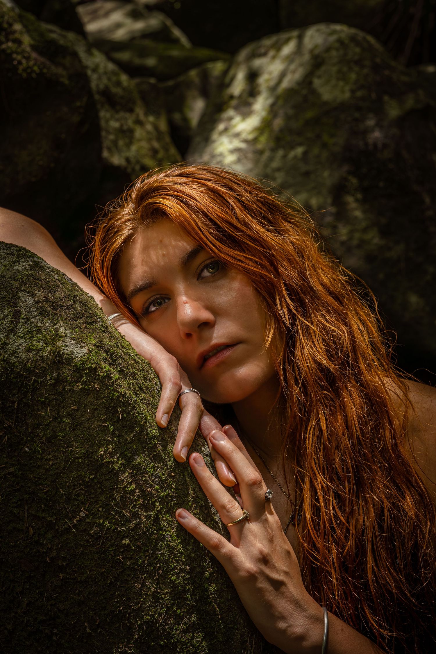 A red-haired woman rests her cheek on a mossy rock, gazing upward with rings on her fingers in a forest setting.