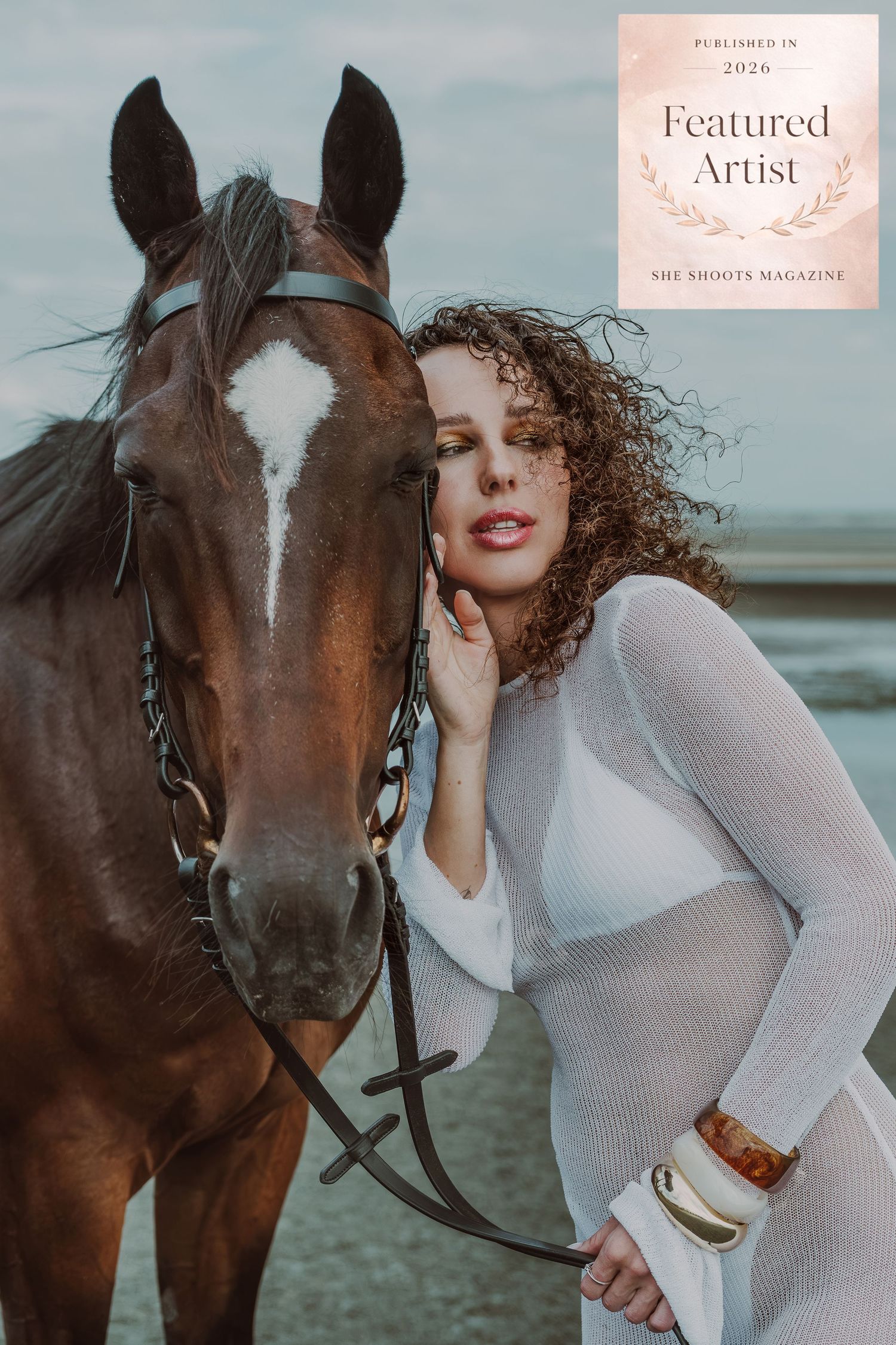A curly-haired woman in a white sheer dress poses closely with a brown horse outdoors, featured in She Shoots Magazine 2026.