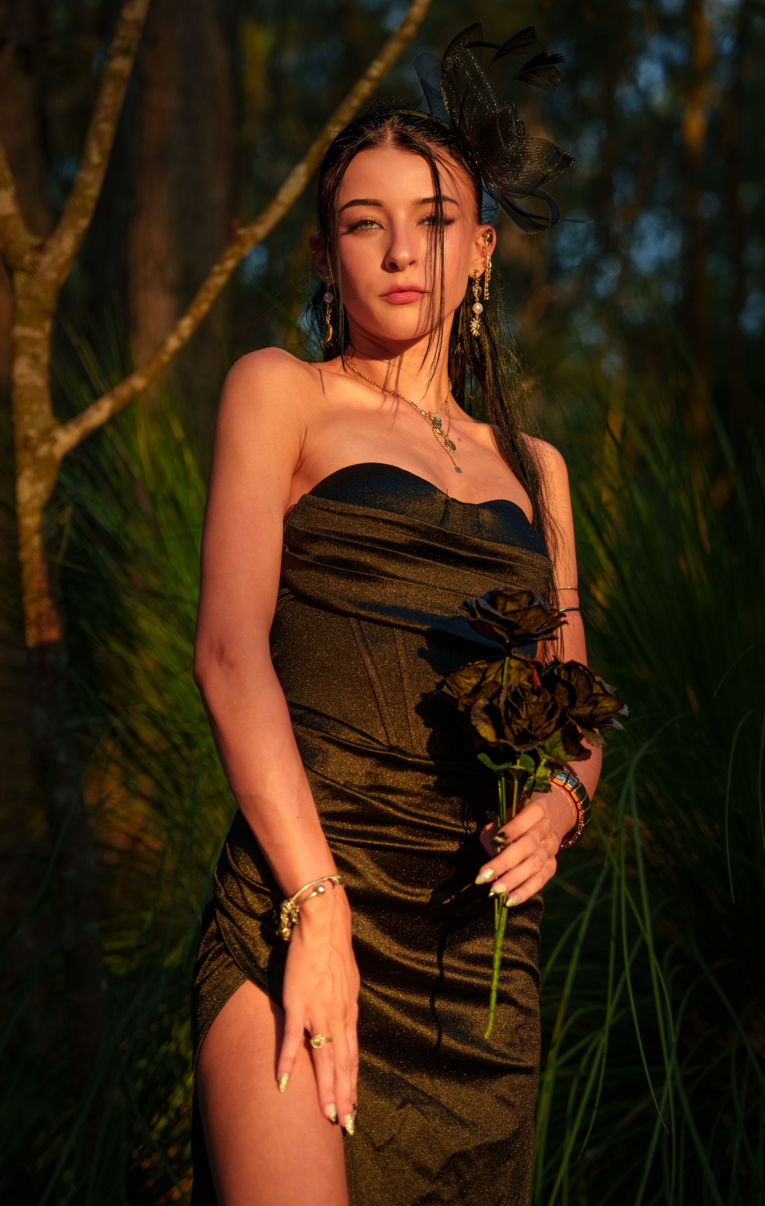 Woman in black strapless dress holding dark roses, wearing fascinator and jewelry, posing outdoors in golden hour light.