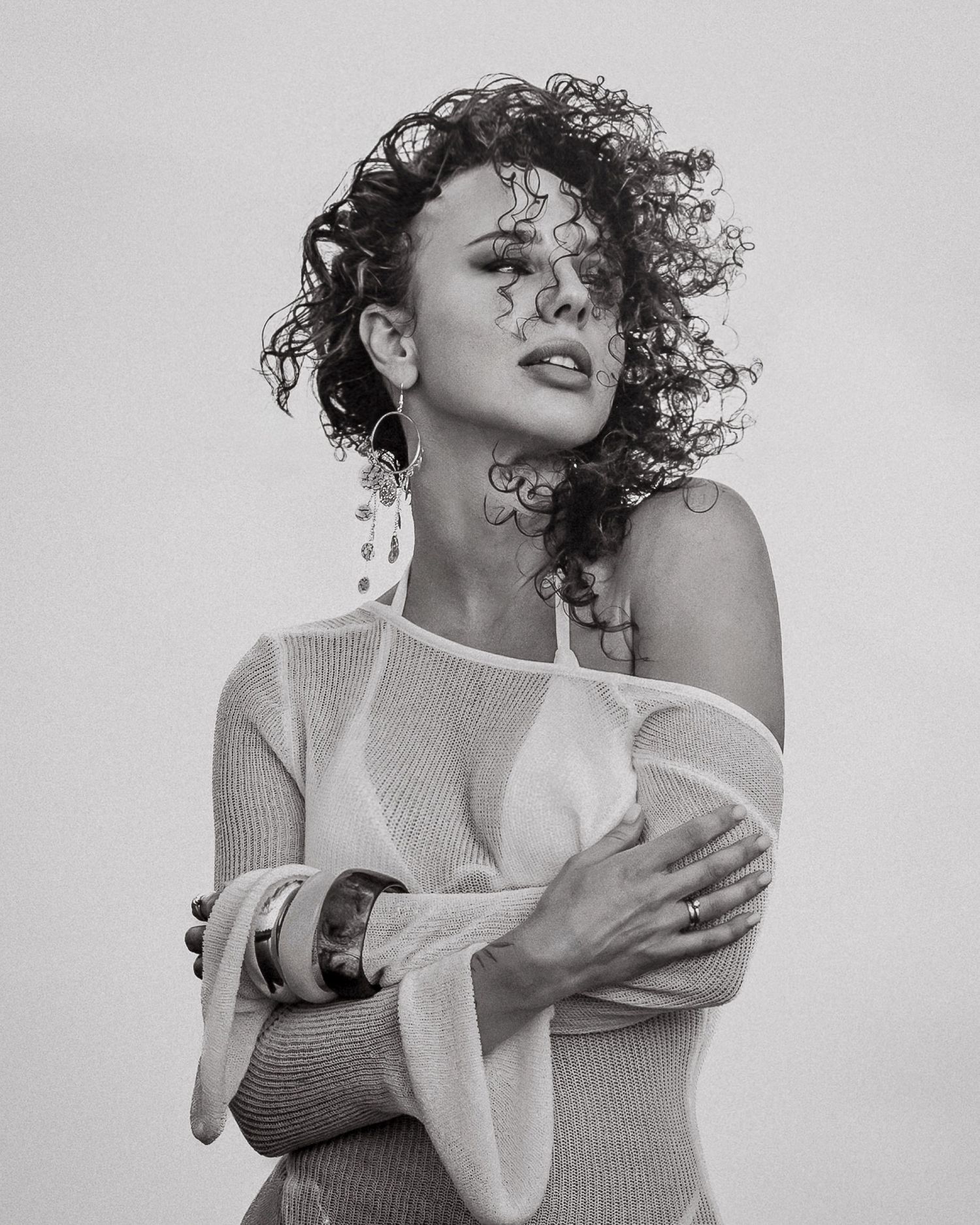 Black and white portrait of a curly-haired woman in off-shoulder top, arms crossed, wearing statement earrings.