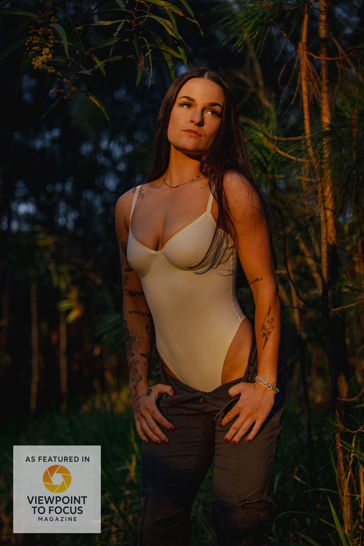 A person poses in a beige bodysuit against a moody forest backdrop during golden hour lighting.