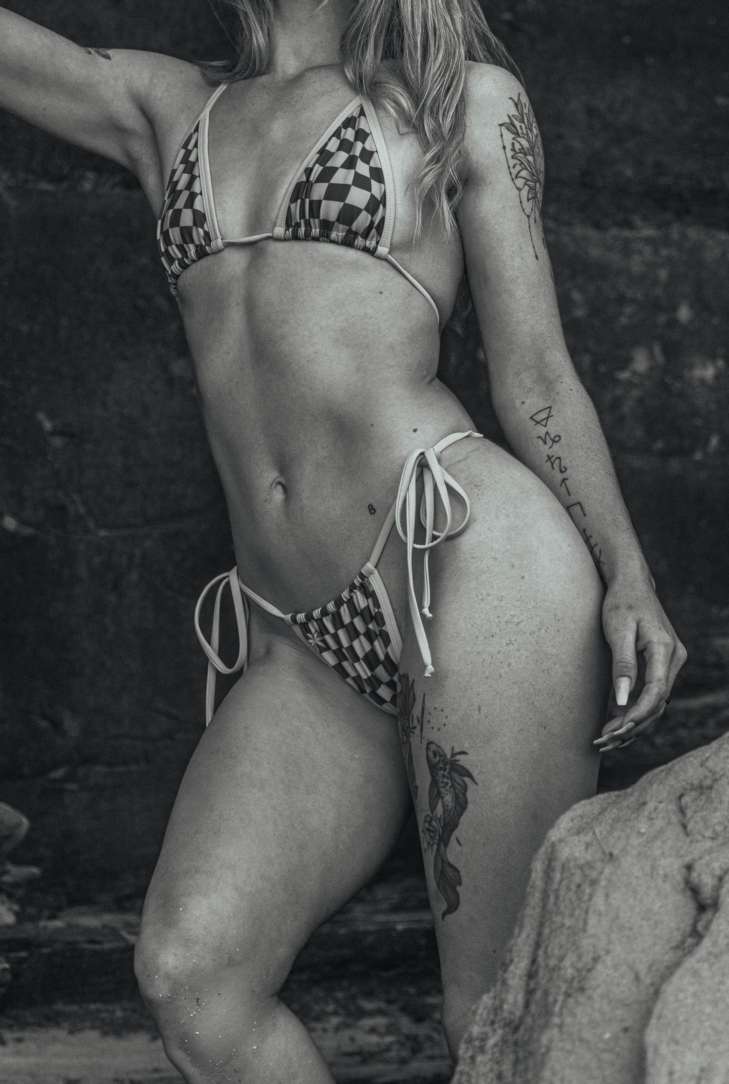 Woman in checkered bikini with tattoos posing on rocky beach in black and white photo.