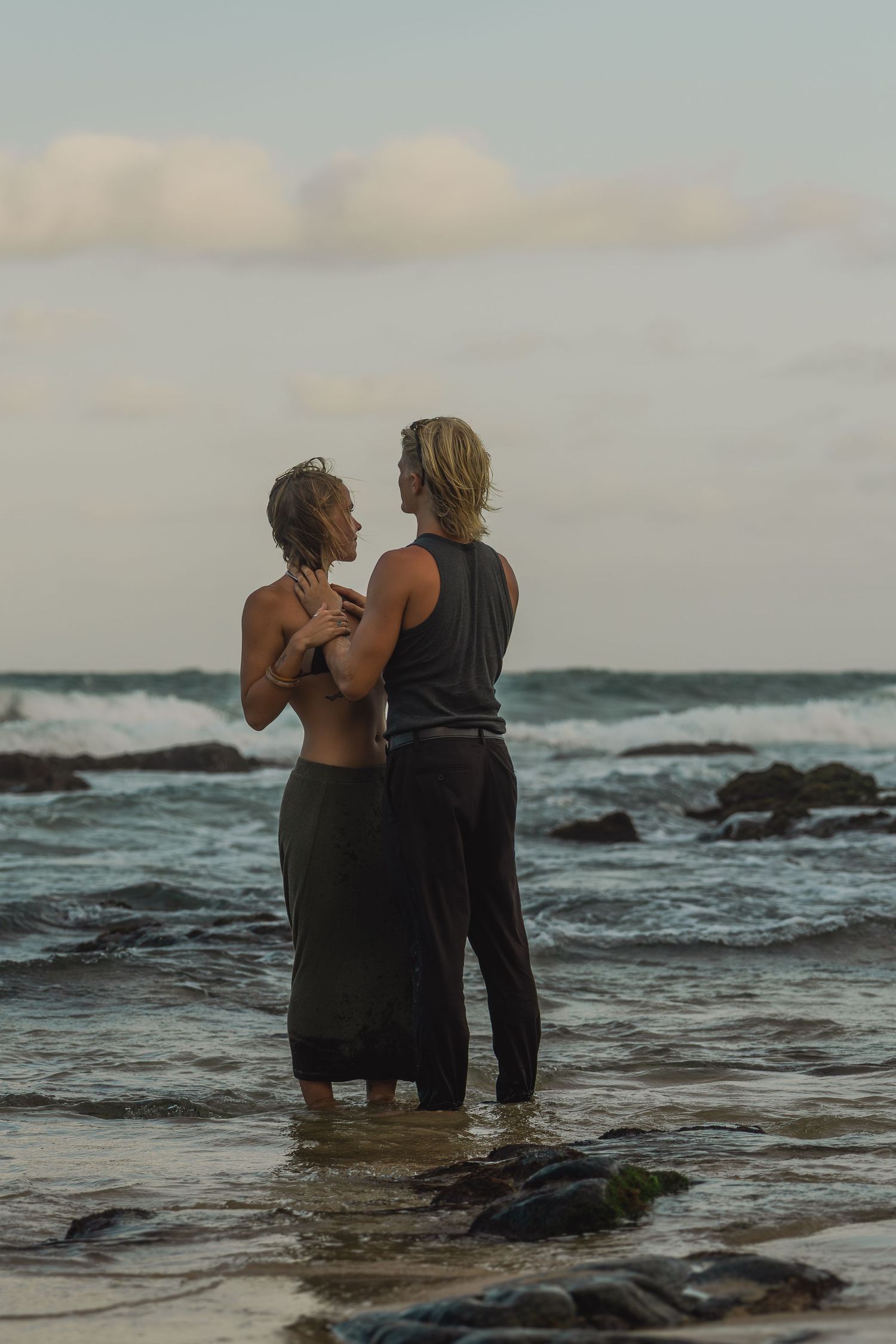 Two people stand together on rocky shore at sunset as ocean waves crash behind them.