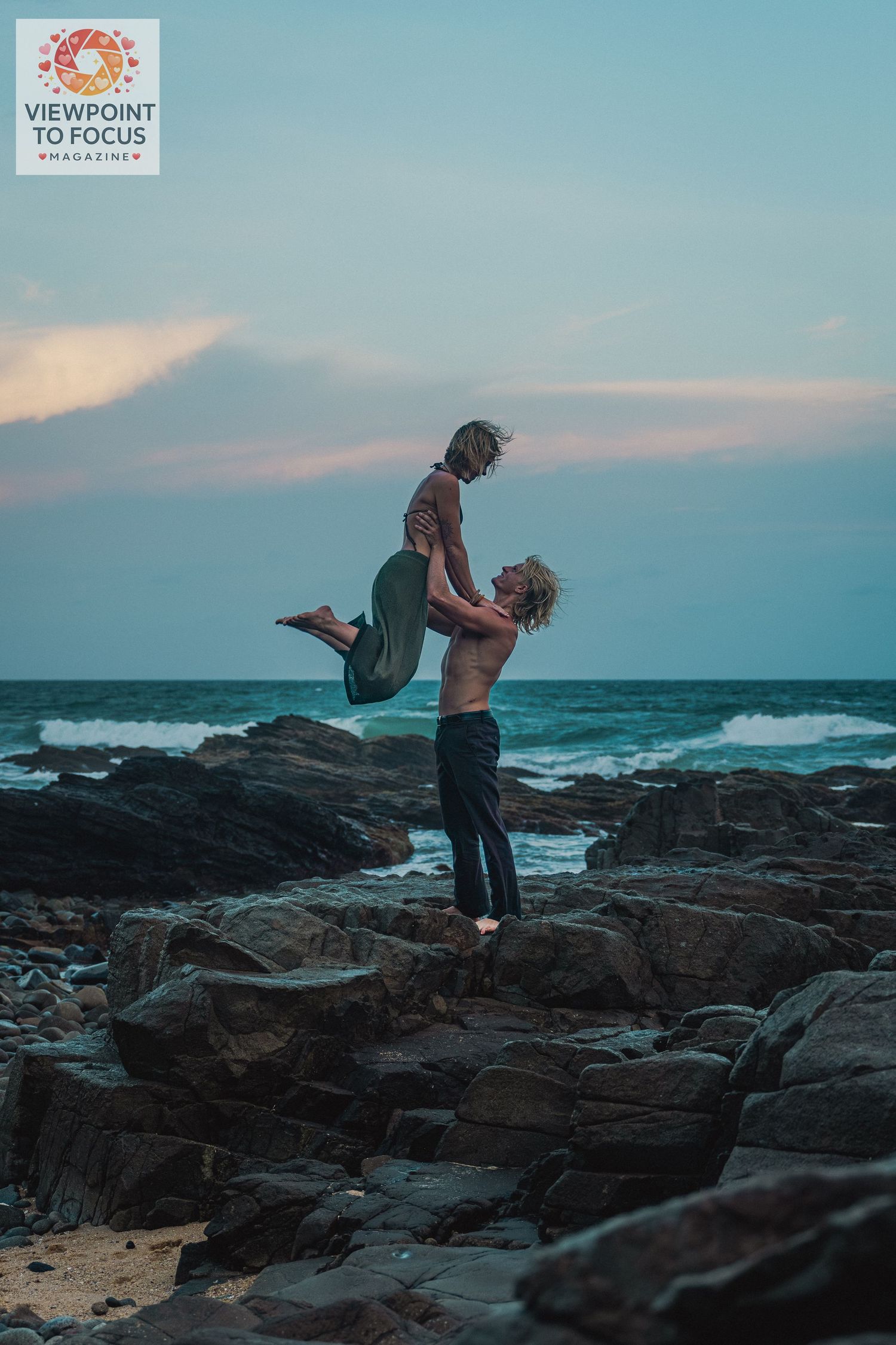 A man lifts a woman overhead on rocky coastal shore at dusk with ocean waves in the background.