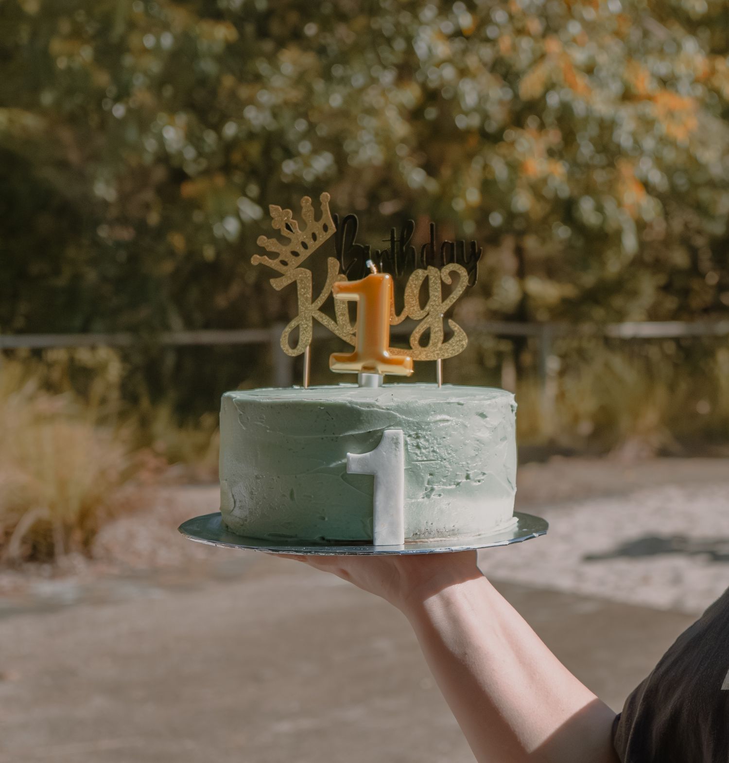 A light blue birthday cake being held up outdoors against a natural background.