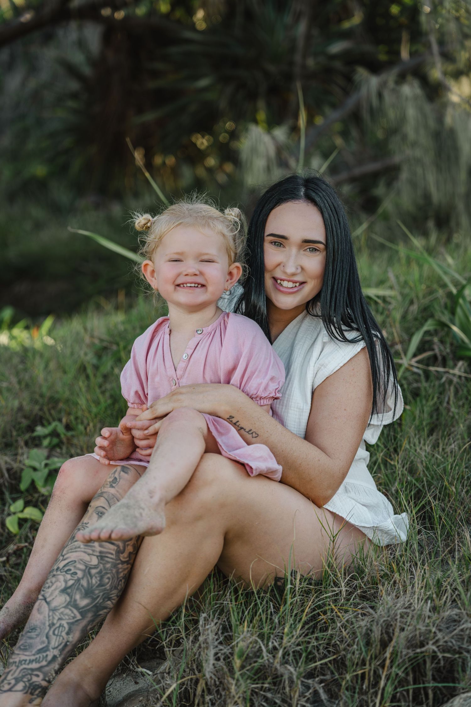 A smiling dark-haired woman sits outdoors in grass holding a laughing blonde toddler girl in a pink dress.