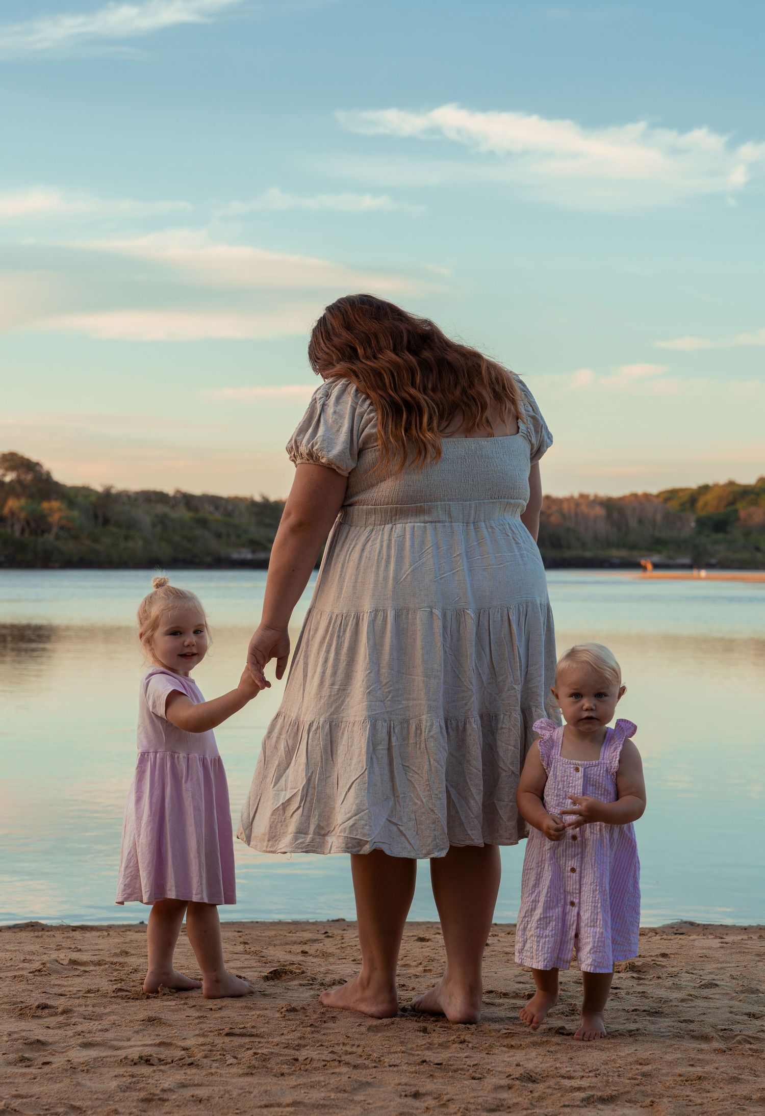 Mother and two small children standing barefoot on beach at sunset.
