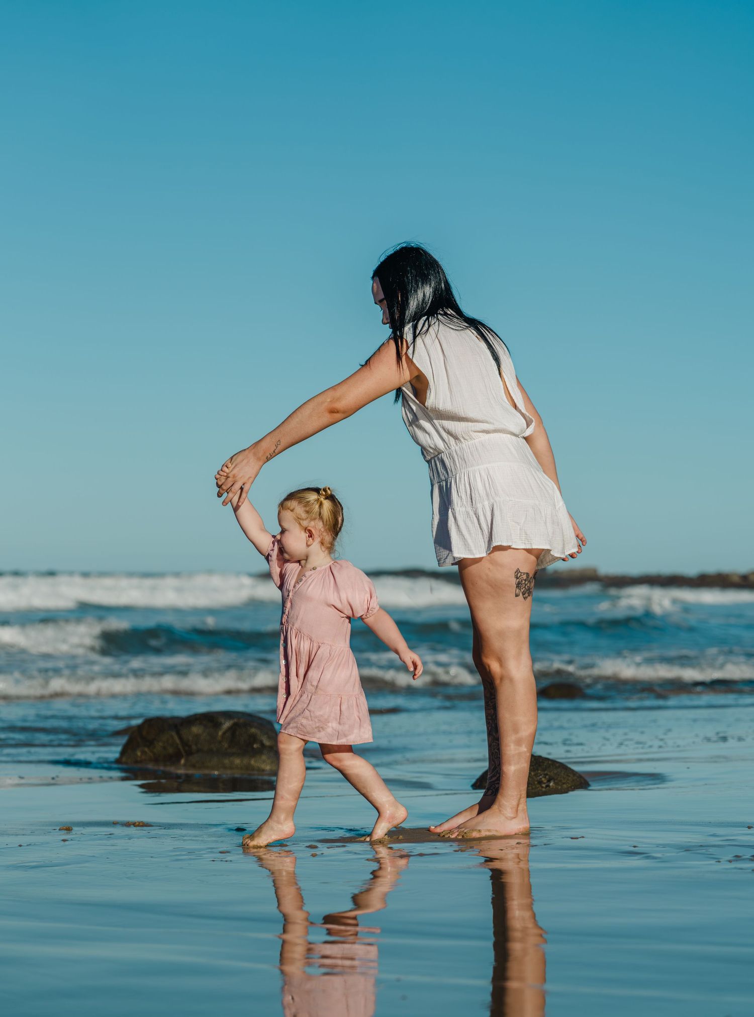 A mother and young daughter dance barefoot on a reflective beach with ocean waves and clear blue sky.