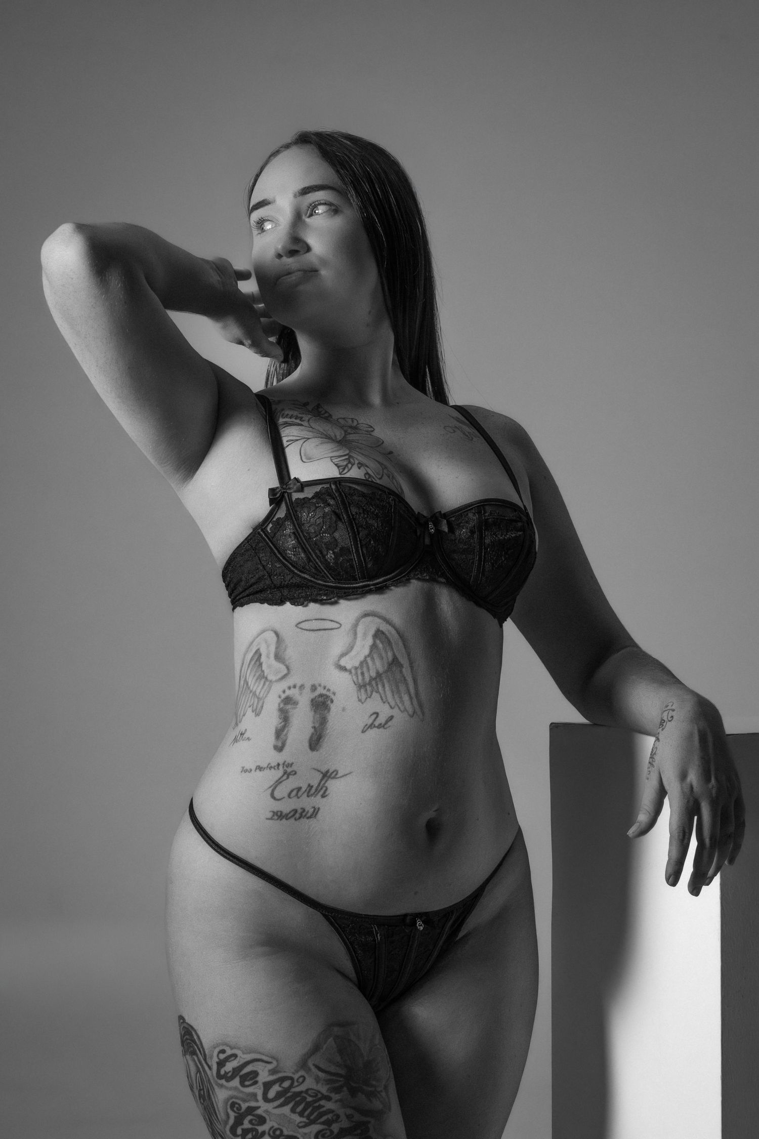 Black and white boudoir photo showing a person in lingerie with visible tattoos posing against a plain background.