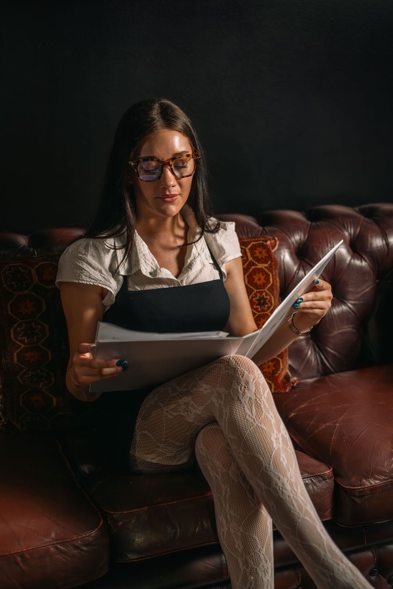 Person in glasses and lace stockings reads documents while sitting on leather couch.