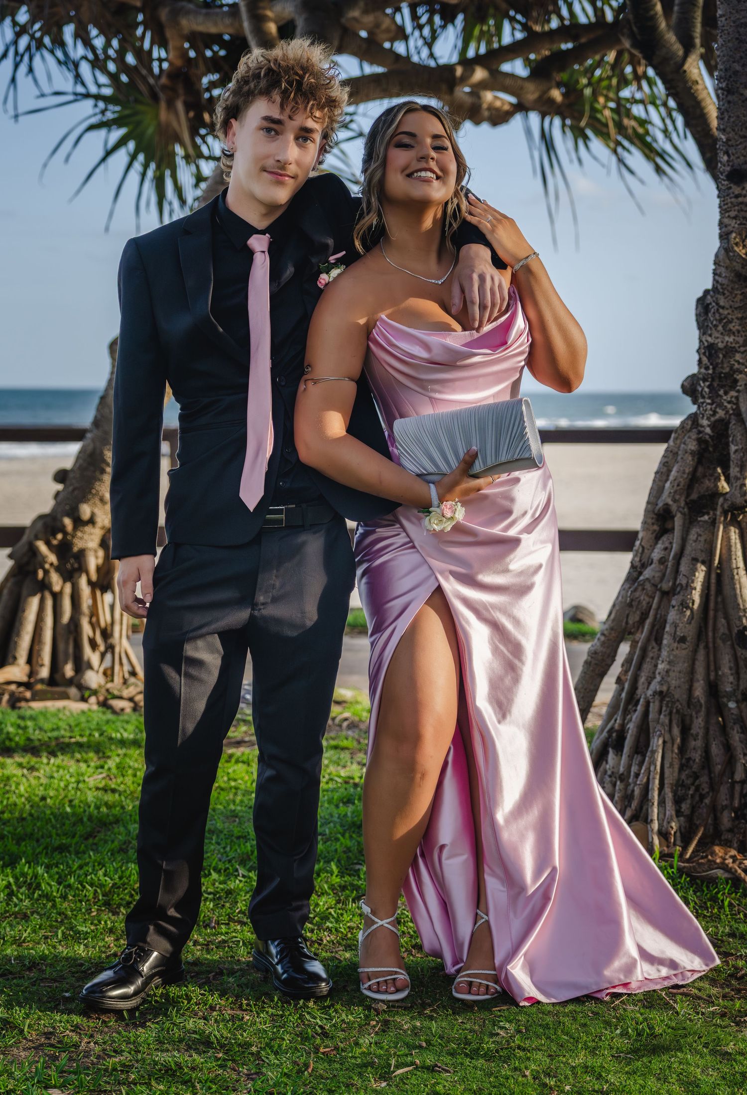 Two people in formal attire share a candid moment by palm trees on a beachfront.