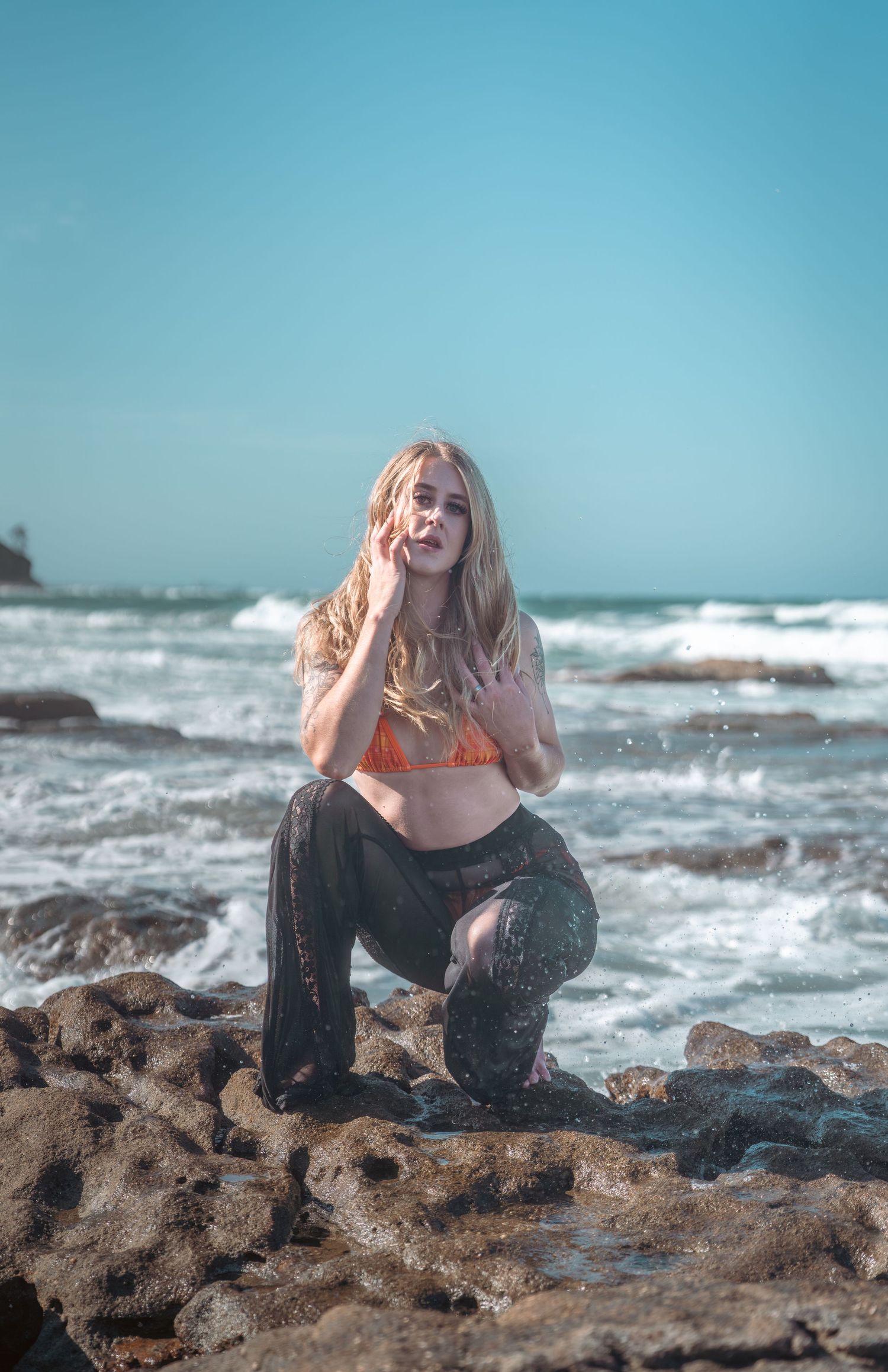 Blonde woman in orange bikini top and black pants posing on rocky beach with ocean waves behind her.