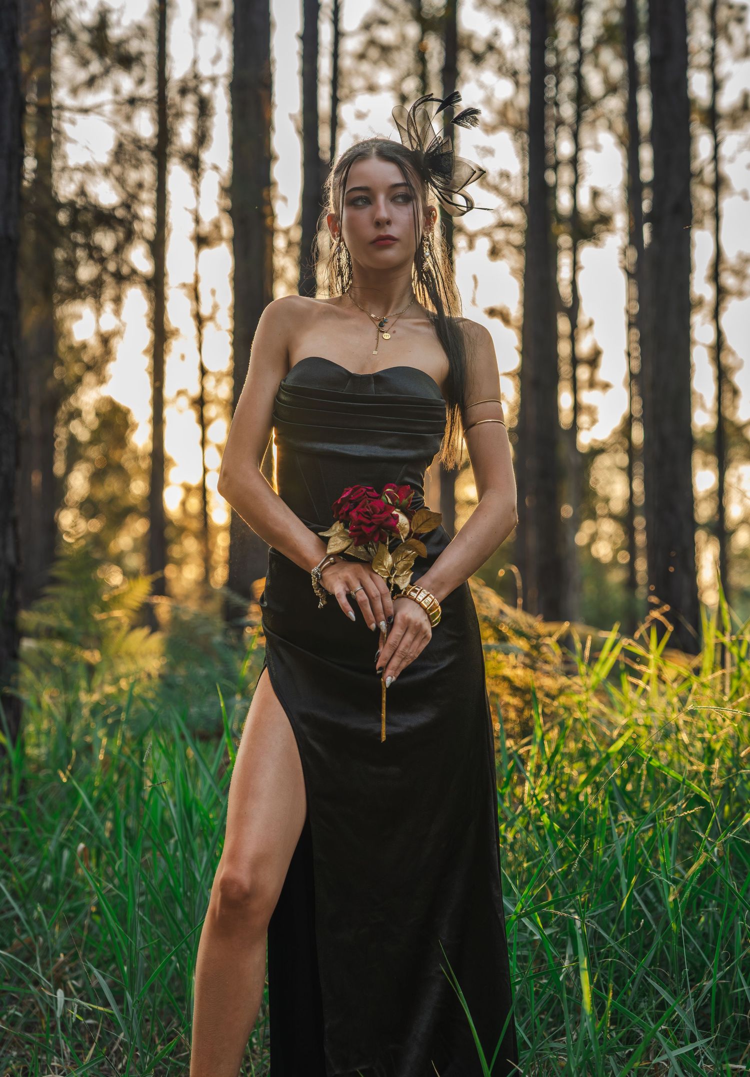 Woman in black strapless gown holding red roses in a sunlit forest, wearing feather hair accessory and gold jewelry.