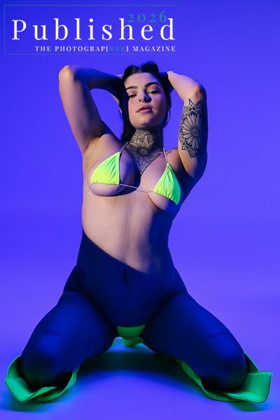 Tattooed woman poses in neon green bikini top and blue pants under purple studio lighting for Published photography magazine.
