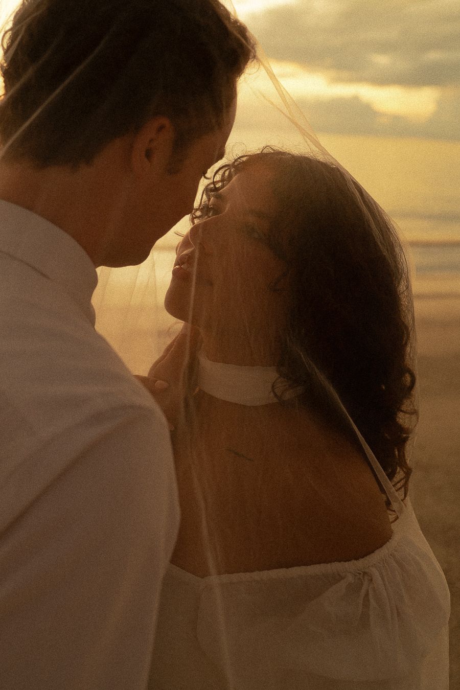 Romantic couple sharing an intimate moment on a beach at golden sunset, bride in white dress with veil.