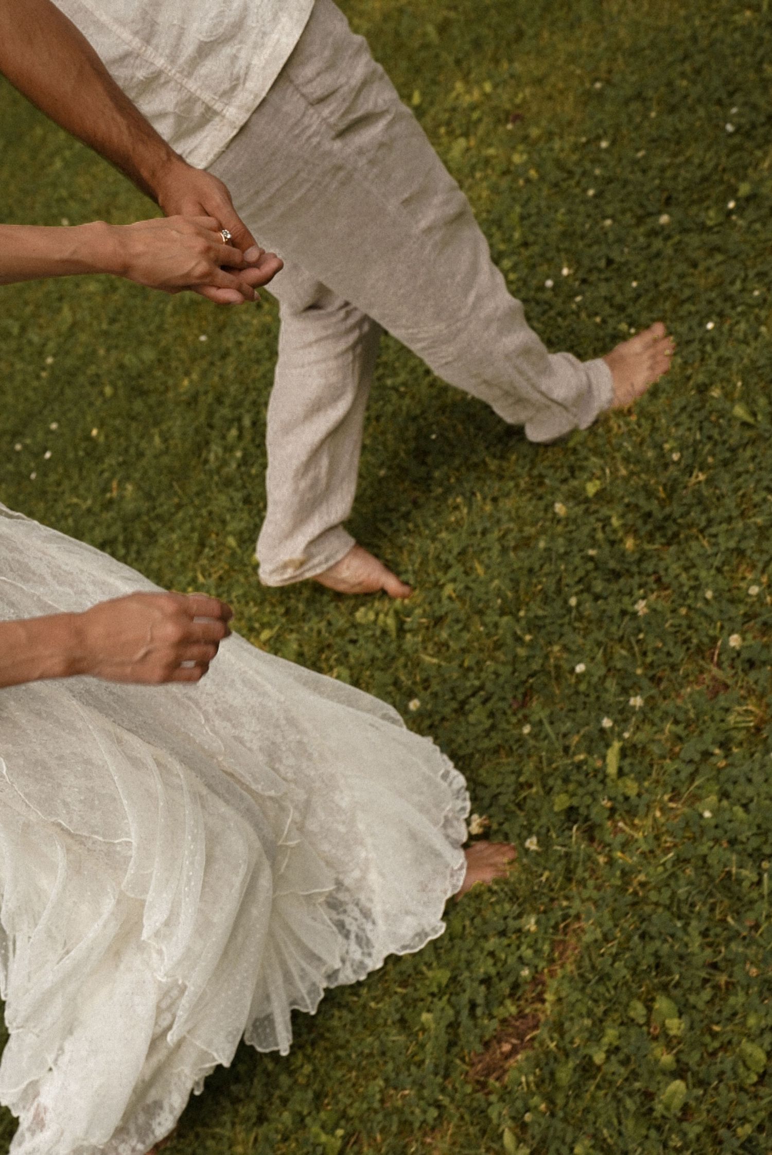 Barefoot couple in white attire holding hands on lush green grass, bride's lace dress spread elegantly.