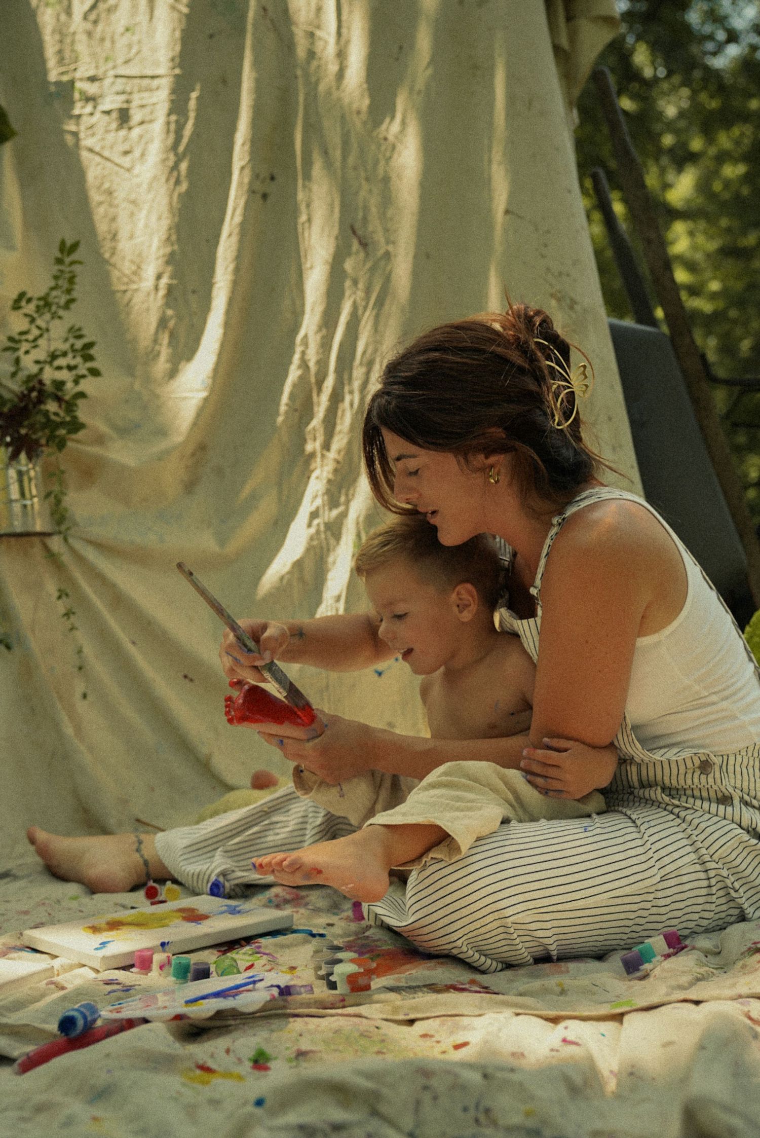 A mother and toddler sit together under a canvas tent, painting with colorful supplies on a sunny day.