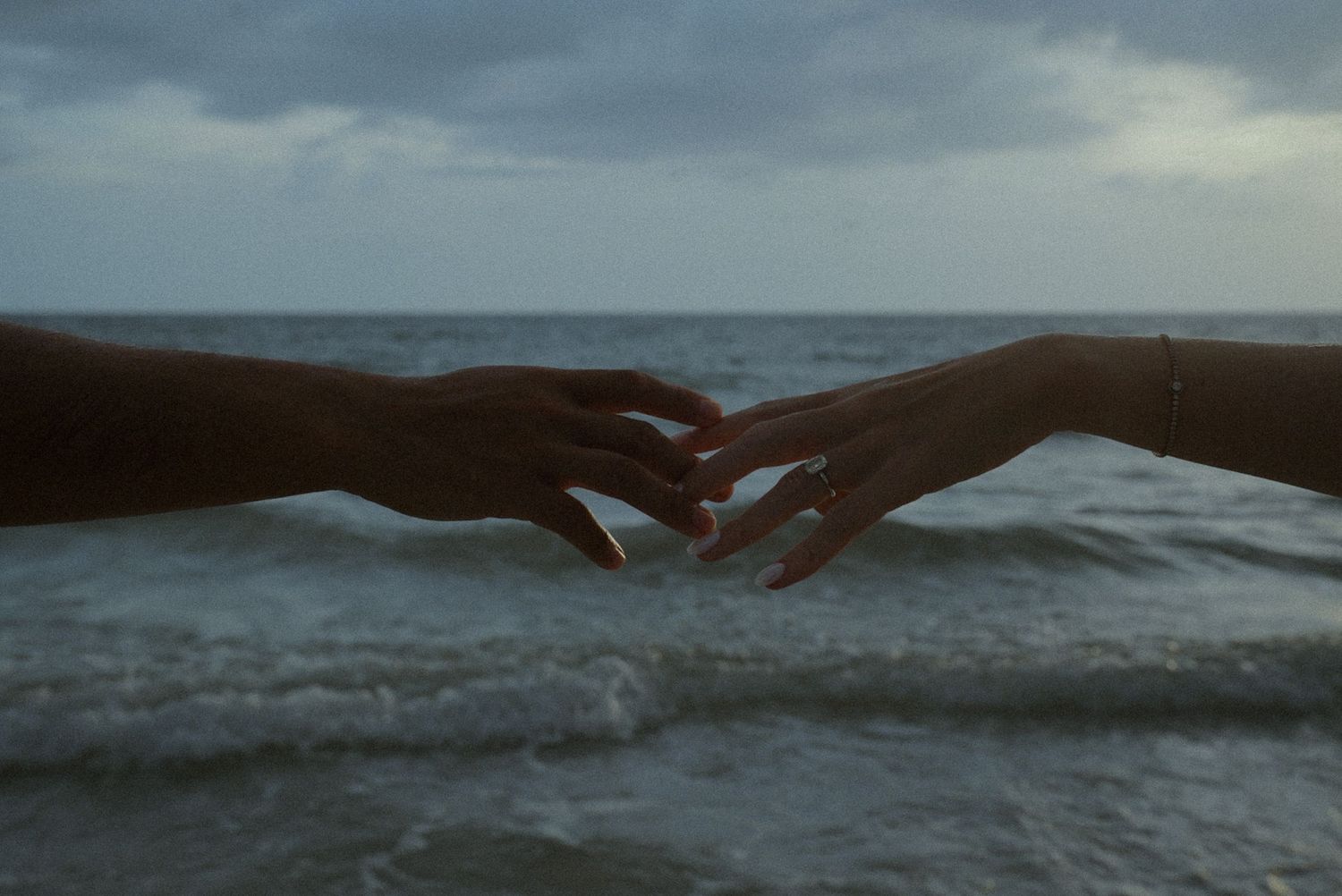 Two hands reaching toward each other over ocean waves at dusk, one wearing a bracelet, in a romantic beach scene.