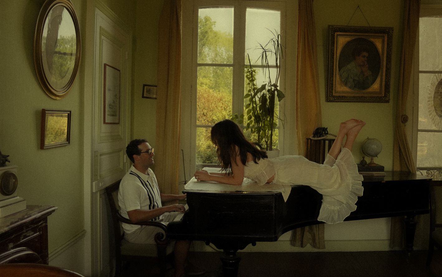 A man sits at a desk while a woman reclines on a bed near a sunlit window in an elegant green room.