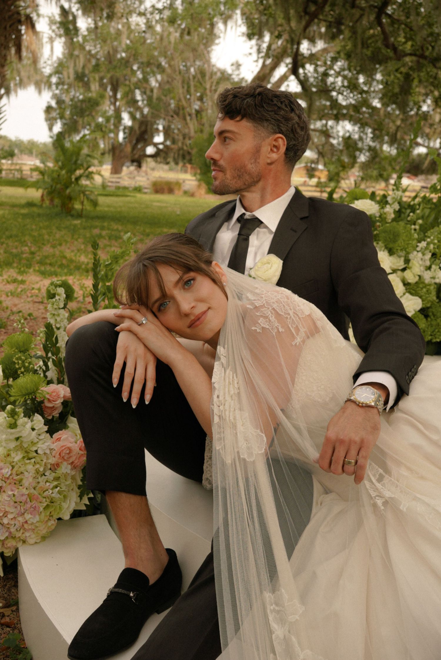 A bride in lace veil leans on groom in black suit amid floral arrangements outdoors under moss-draped trees.