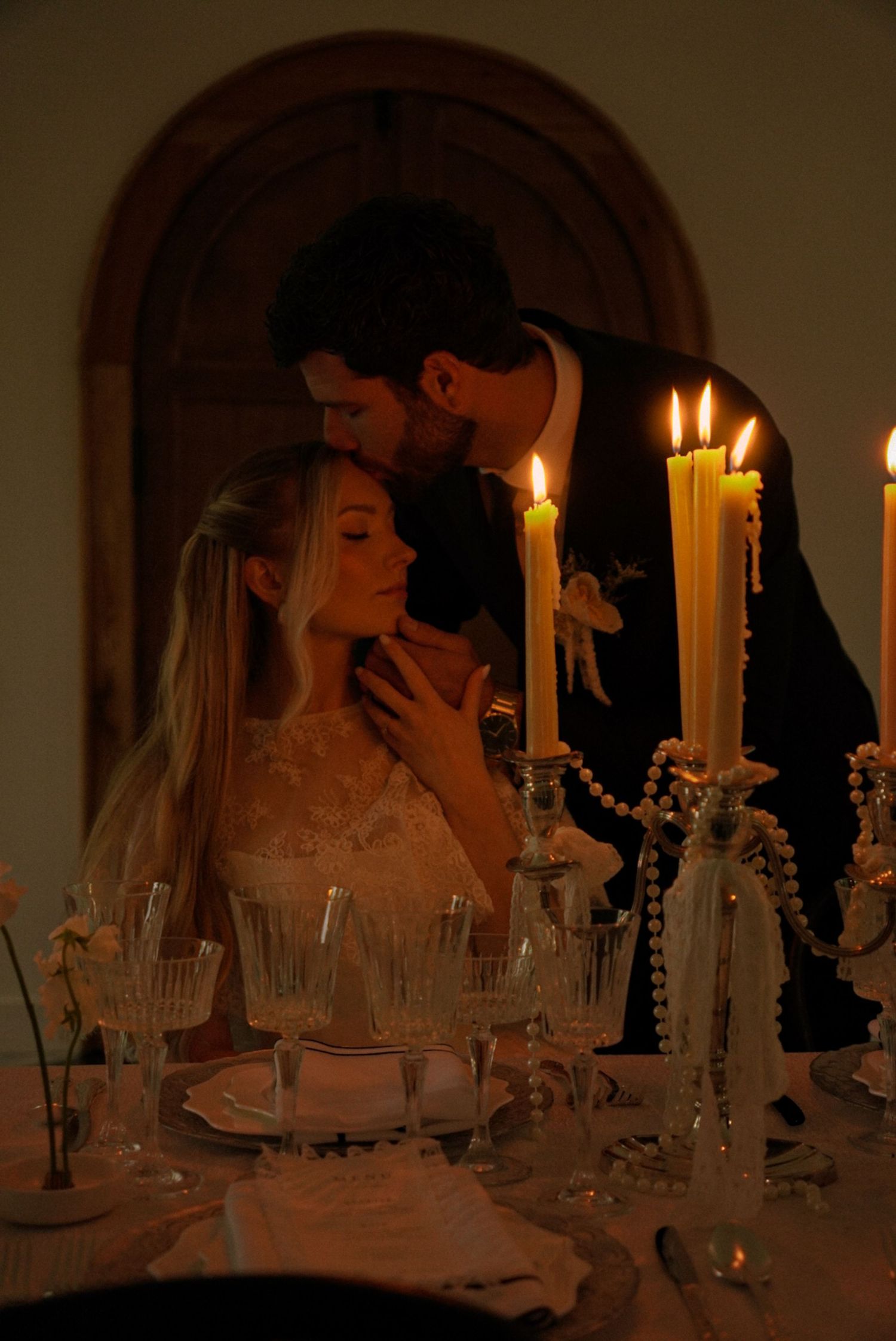 Romantic candlelit wedding couple sharing intimate moment at elegant reception table with crystal glassware.