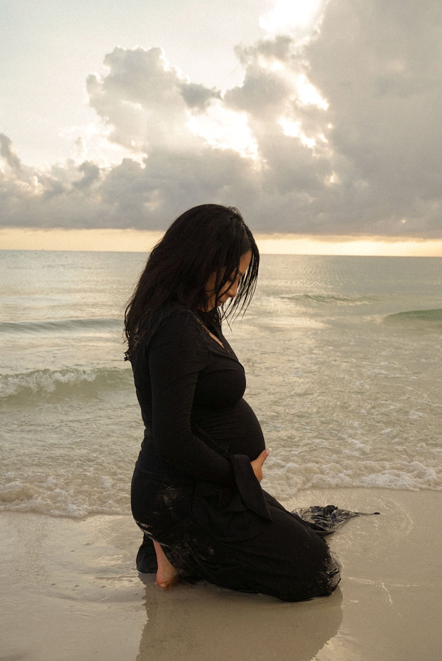 Pregnant woman in black dress kneeling on sandy beach at sunset with ocean waves and cloudy sky.