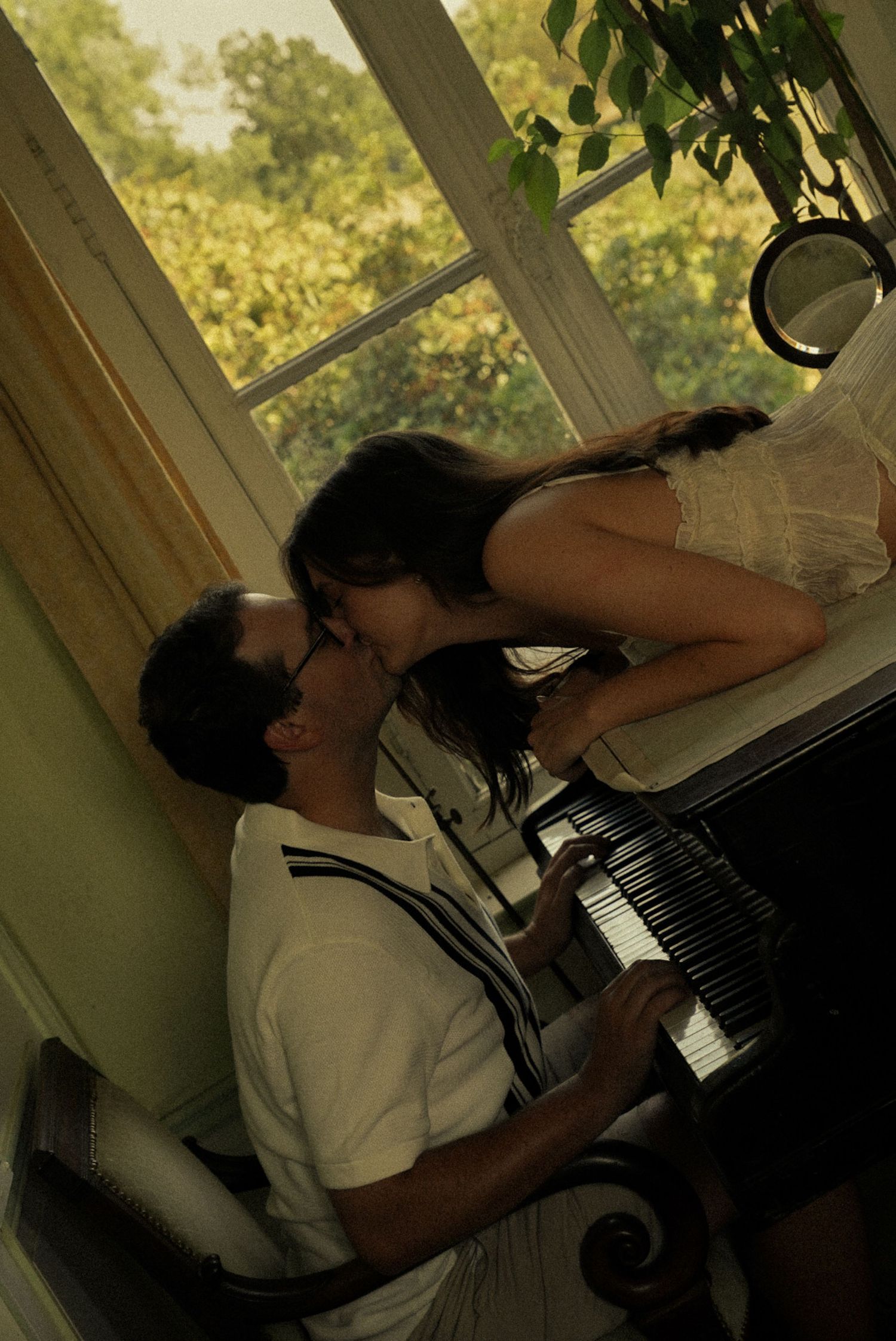 A couple shares a romantic kiss near a piano by a sunlit window with lush green plants in the background.