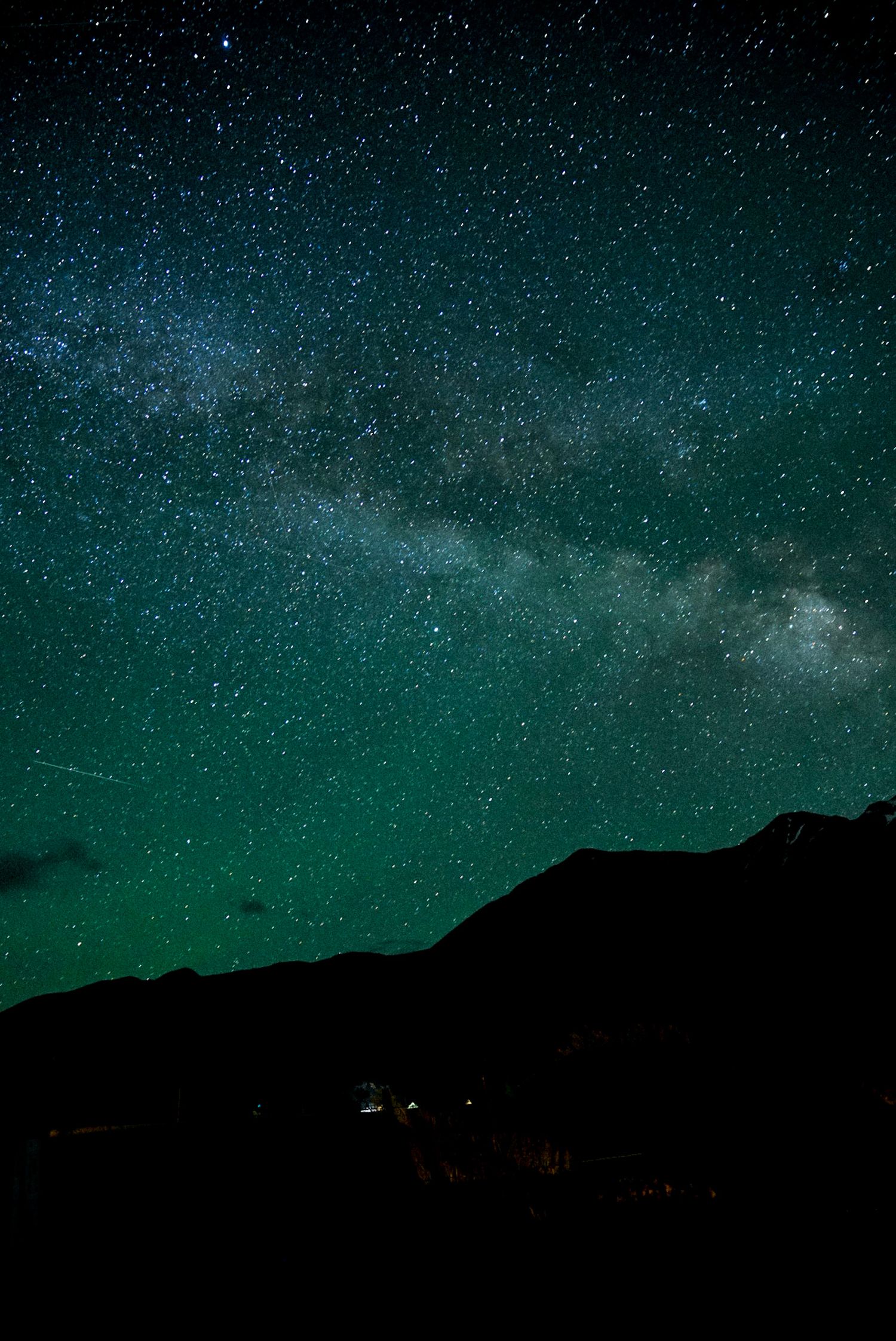 Milky Way galaxy arching over dark mountain silhouette under a stunning green-tinted starry night sky.