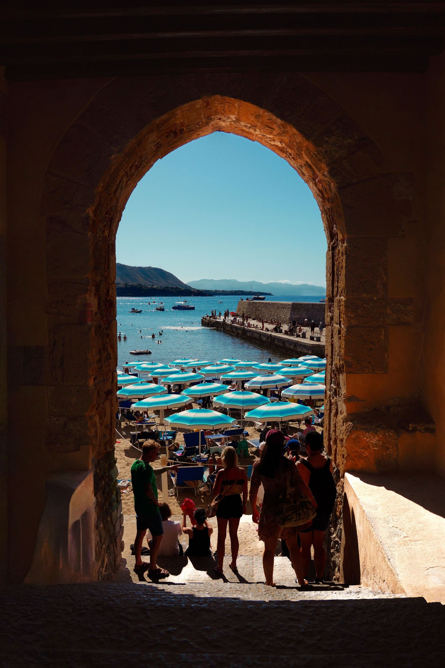 View through stone archway of crowded beach with blue umbrellas and mountains in background.